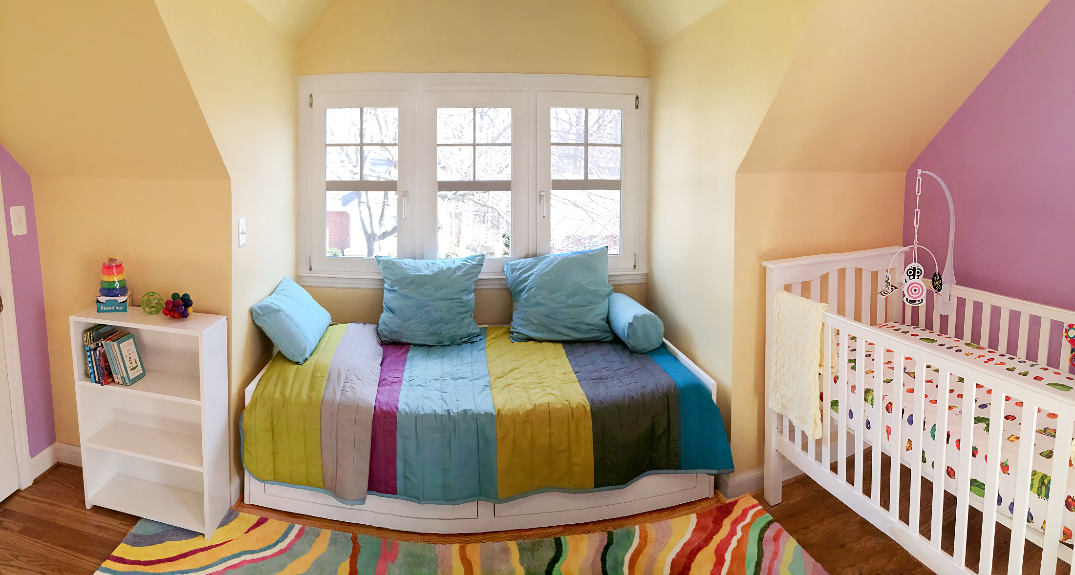 A brightly colored children's bedroom with a rainbow-striped rug, a daybed with rainbow-colored bedding and large blue pillows beneath a window, and a white crib with a colorful bedding and hanging toys on the right. There is a white bookshelf with t