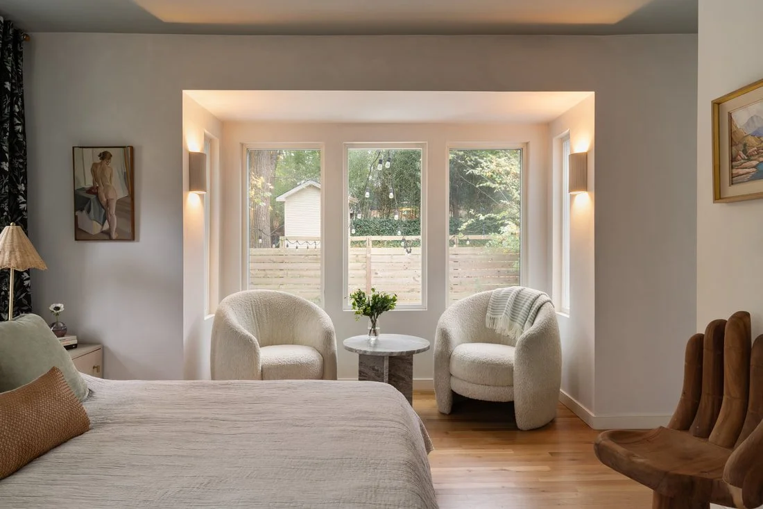 A cozy bedroom sitting area with two white upholstered armchairs, a small round table with a vase of greenery, a bed with neutral bedding, and a bay window with backlighting overlooking a backyard with trees and a wooden fence.