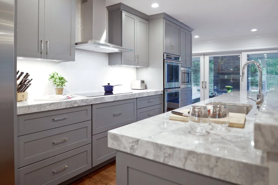 Modern kitchen with gray cabinets, marble countertops, and stainless steel appliances. There are two glasses, a cutting board, and a sink on the island counter, with a large window in the background.