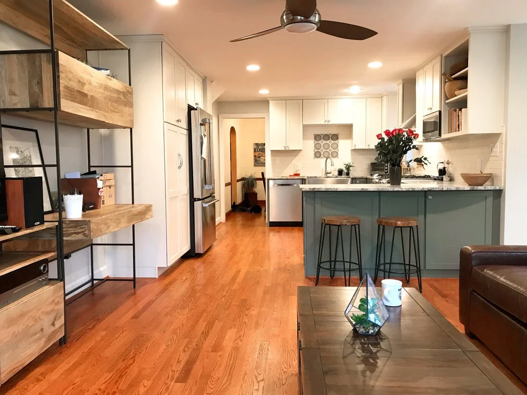 Open-concept kitchen and living room with hardwood floors, white cabinets, a kitchen island with bar stools, a ceiling fan, and decorative items such as a bouquet of red roses and a terrarium on the coffee table.
