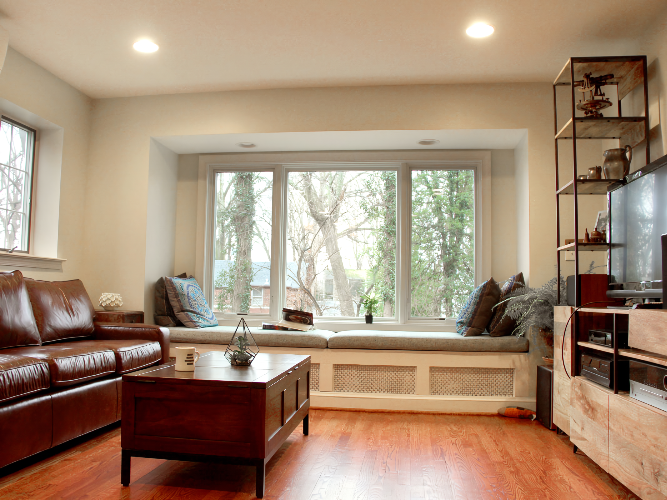 Living room with large window seat, brown leather sofa, wooden coffee table, and a shelving unit with decorative items and a TV.