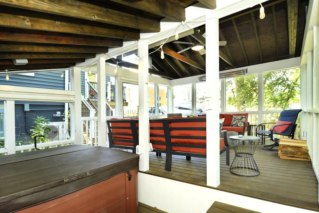 A screened-in porch with wooden ceiling beams, string lights, and outdoor furniture including chairs and a table, overlooking a backyard with trees.