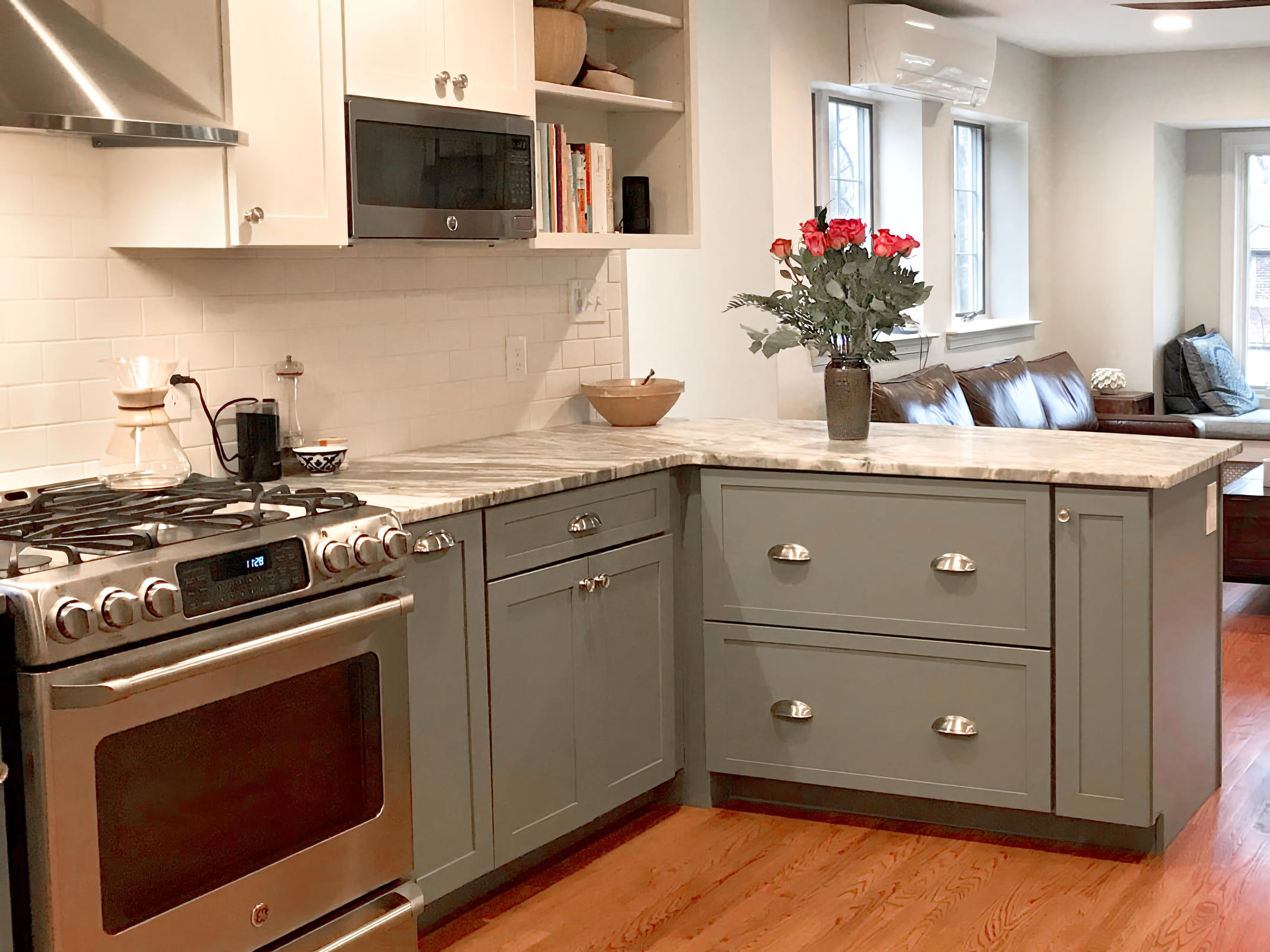 A kitchen with stainless steel oven, white tiles, white upper cabinets, green lower cabinets, marble countertop, and a vase of red roses, with a living room visible in the background.