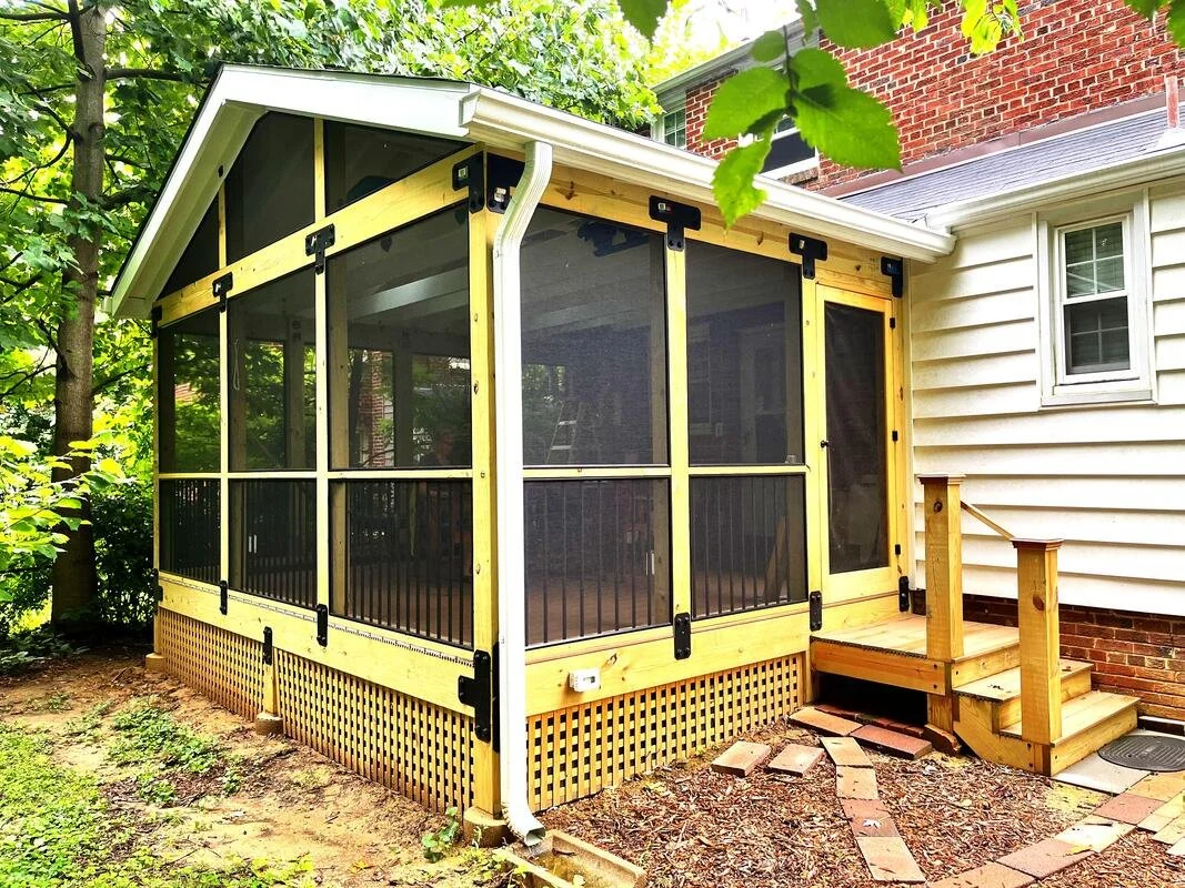 A screened-in porch with wooden framing, attached to a white house, featuring steps leading up to a small deck and surrounded by trees and greenery.