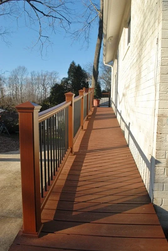 Wooden porch deck with black metal railing attached to a white brick house, under a clear blue sky with leafless trees in the background.