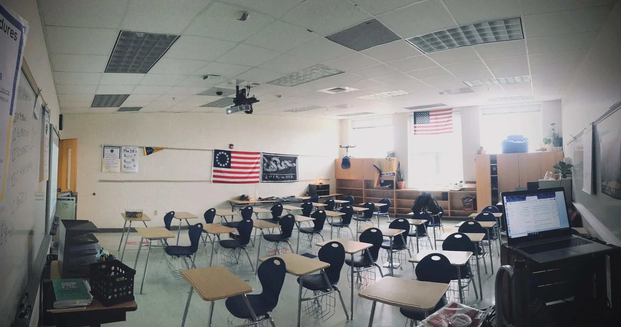 Classroom with student desks and chairs arranged facing the front. American flags and educational posters on the walls, laptop and teaching materials on a desk, sunlight coming through windows at the back.
