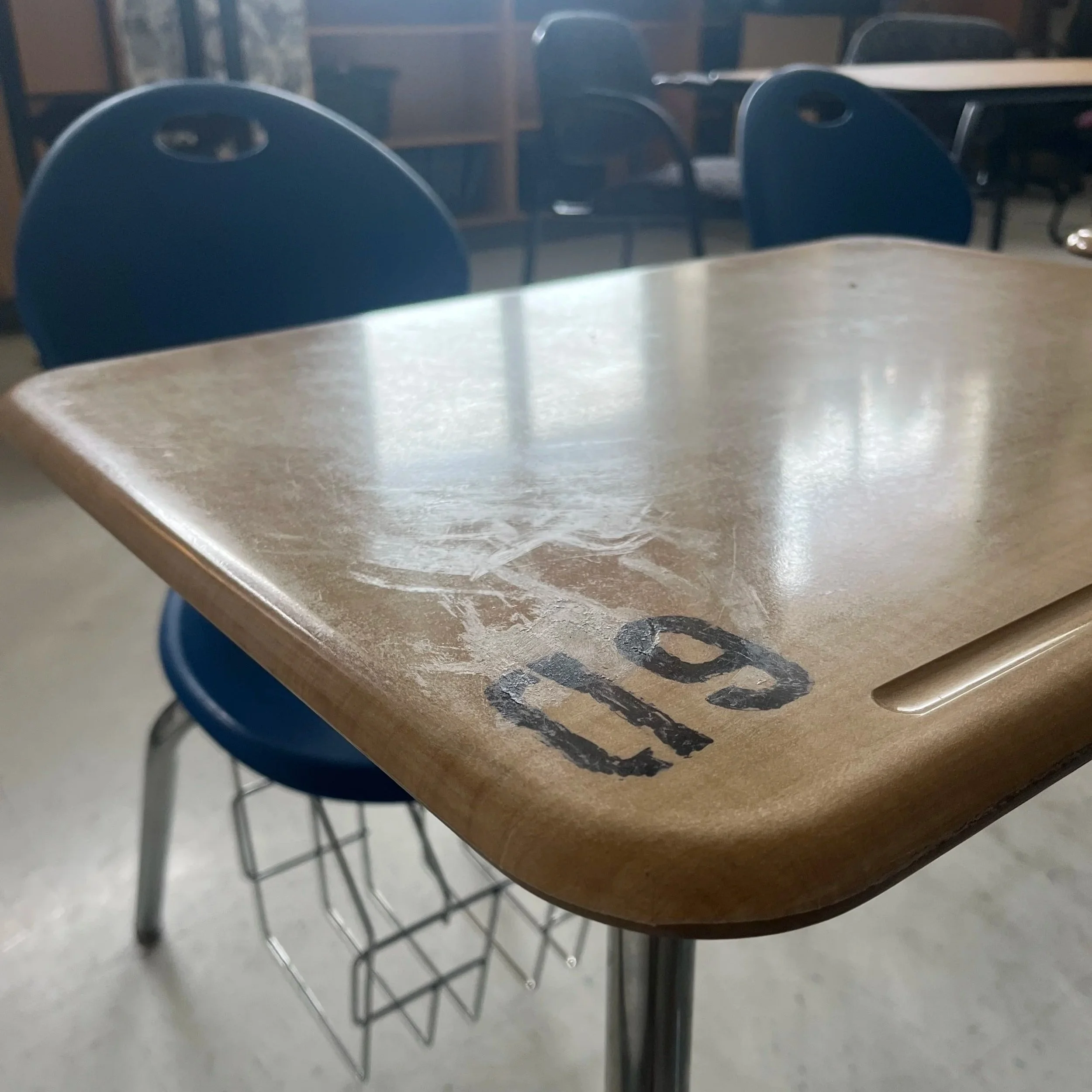 Close-up of a wooden school desk with the number '10' stenciled on the corner, surrounded by blue chairs in a classroom.