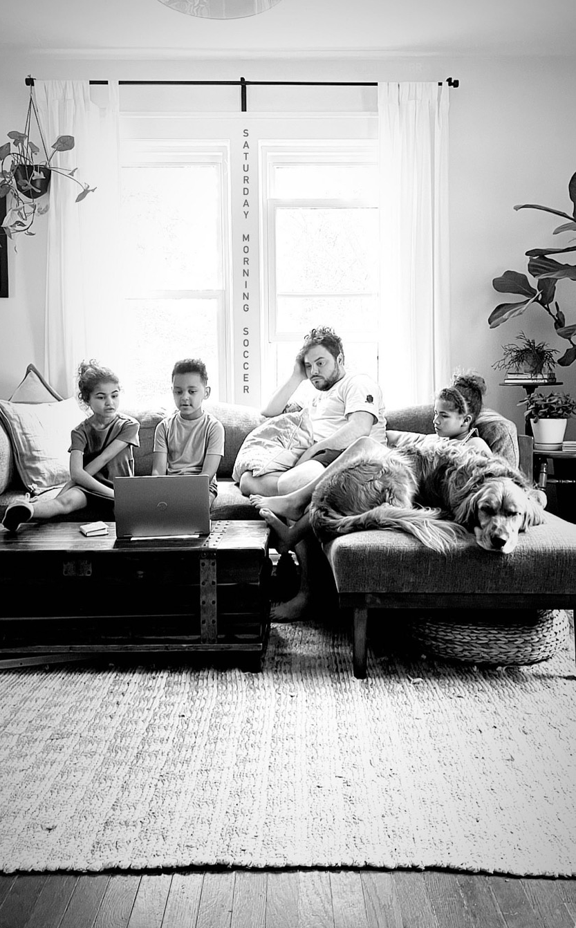A man and three children sitting on a couch in a living room, watching laptop. Two dogs are lying next to the man. Large windows with white curtains in the background, and a sign reading 'Saturday Morning Soccer' between the windows.