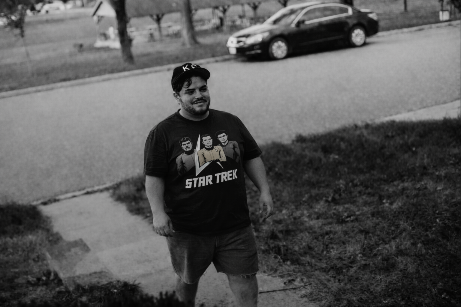 A young man walking on a sidewalk in a residential neighborhood, wearing a Star Trek t-shirt and a black cap with letters KS, with trees and parked cars visible in the background.