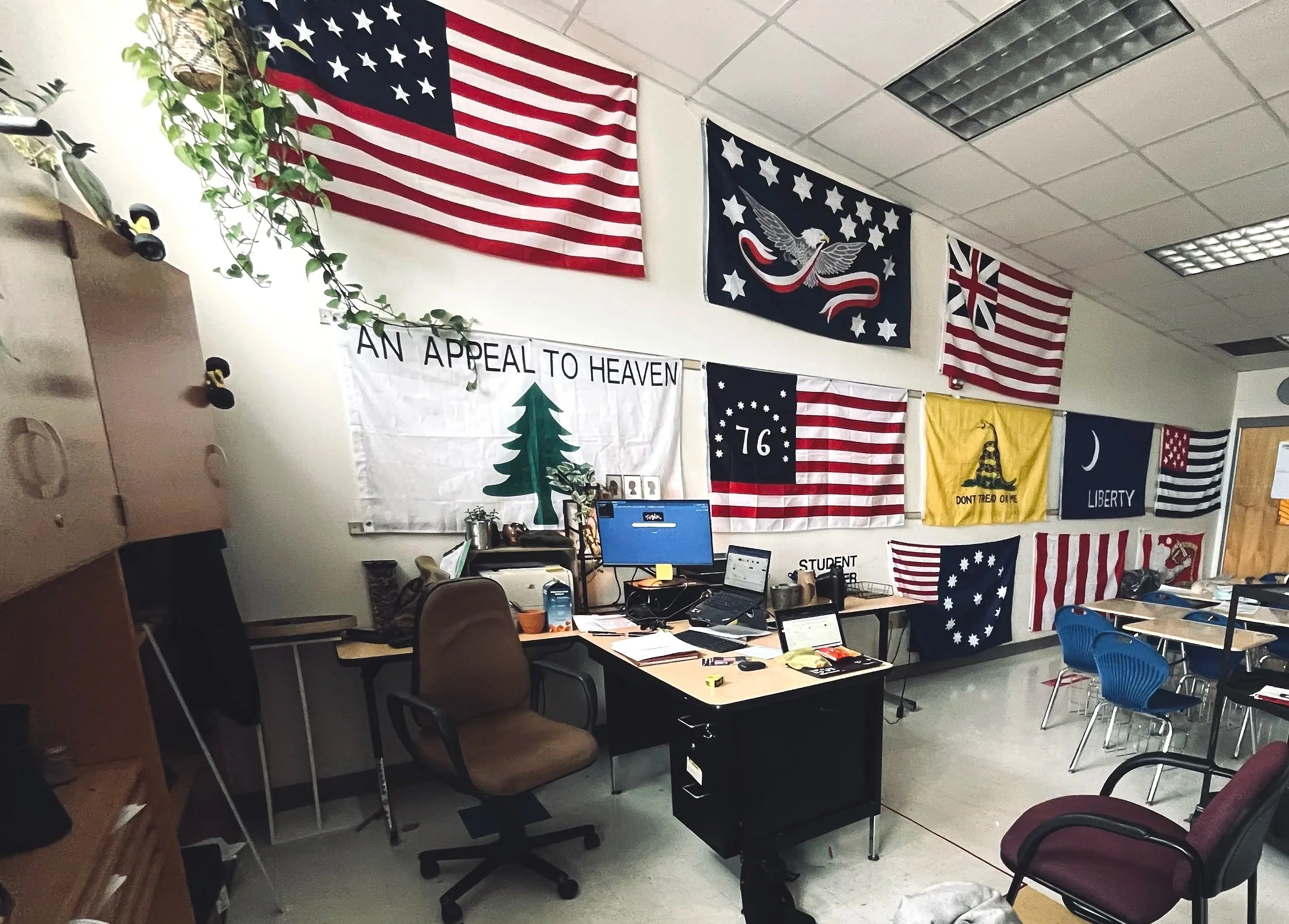 A classroom decorated with various American flags and patriotic banners hanging on the wall, including a large flag, a flag with a bald eagle, and a banner reading 'AN APPEAL TO HEAVEN'. A desk with a computer and office supplies is in the foreground, and chairs are arranged around the room.