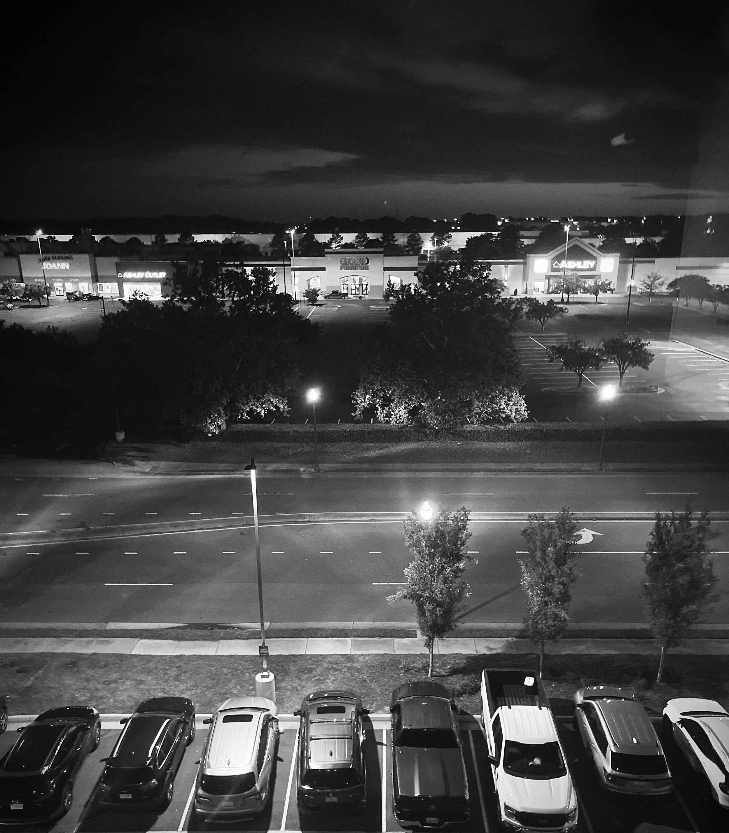 Nighttime view of a parking lot with several cars, trees, and a shopping center in the background, illuminated by streetlights, in black and white.