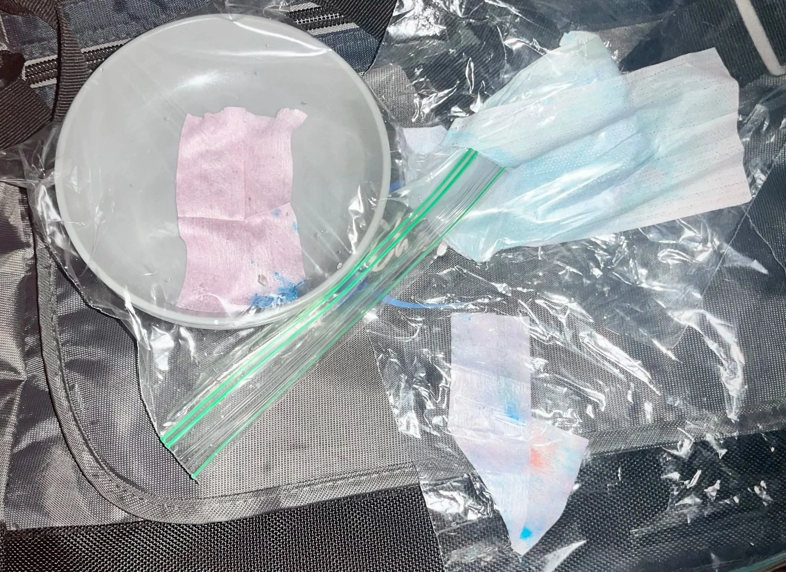 Medical supplies including a pink absorbent pad, a transparent Ziploc bag, an alcohol wipe or pad, and a clear plastic face mask or shield on a textured black surface.