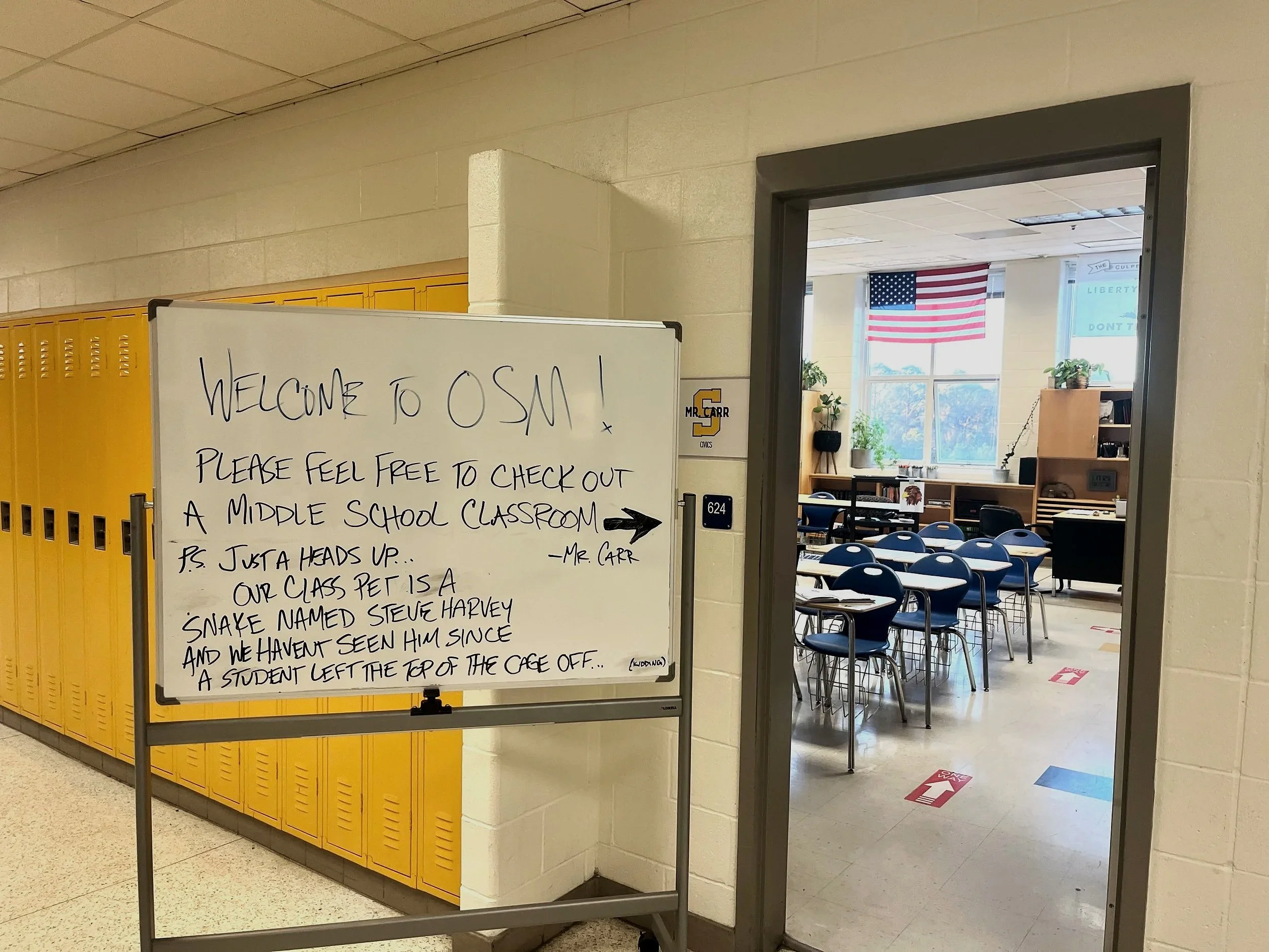 Whiteboard with welcome message outside a middle school classroom, with yellow lockers and classroom visible through the door, American flag in the window.