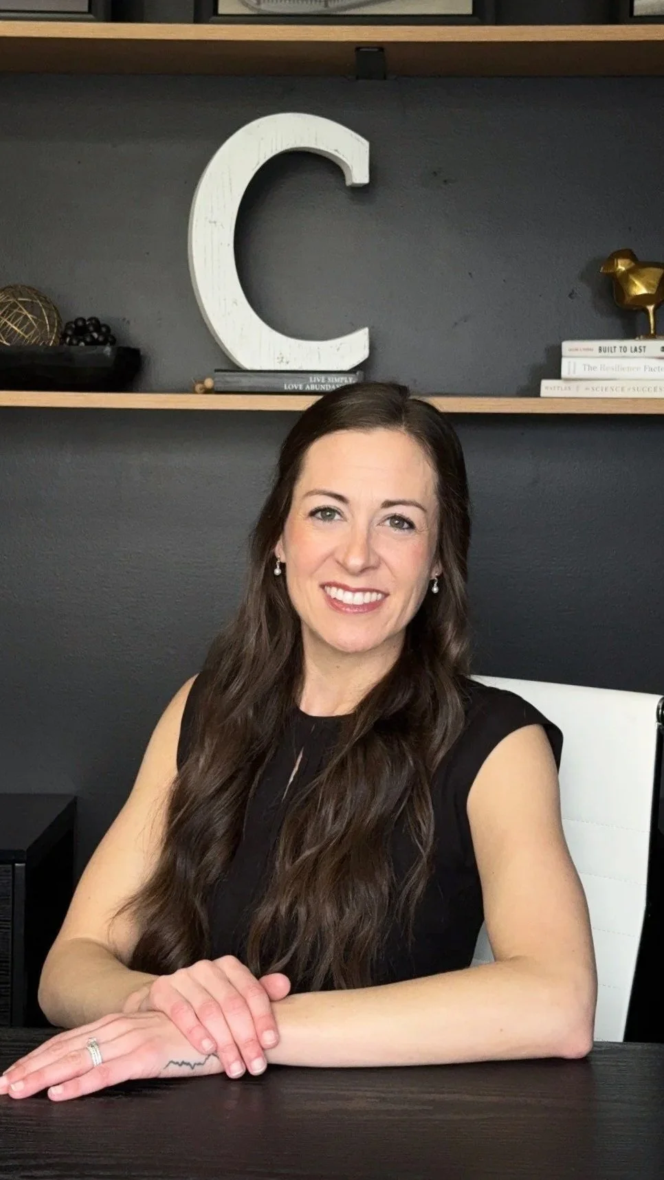 A woman with long dark hair and earrings, wearing a black sleeveless top, sitting at a desk with a smile. Behind her, a large white letter 'C' on a dark wall, with shelves holding books and decorative items.