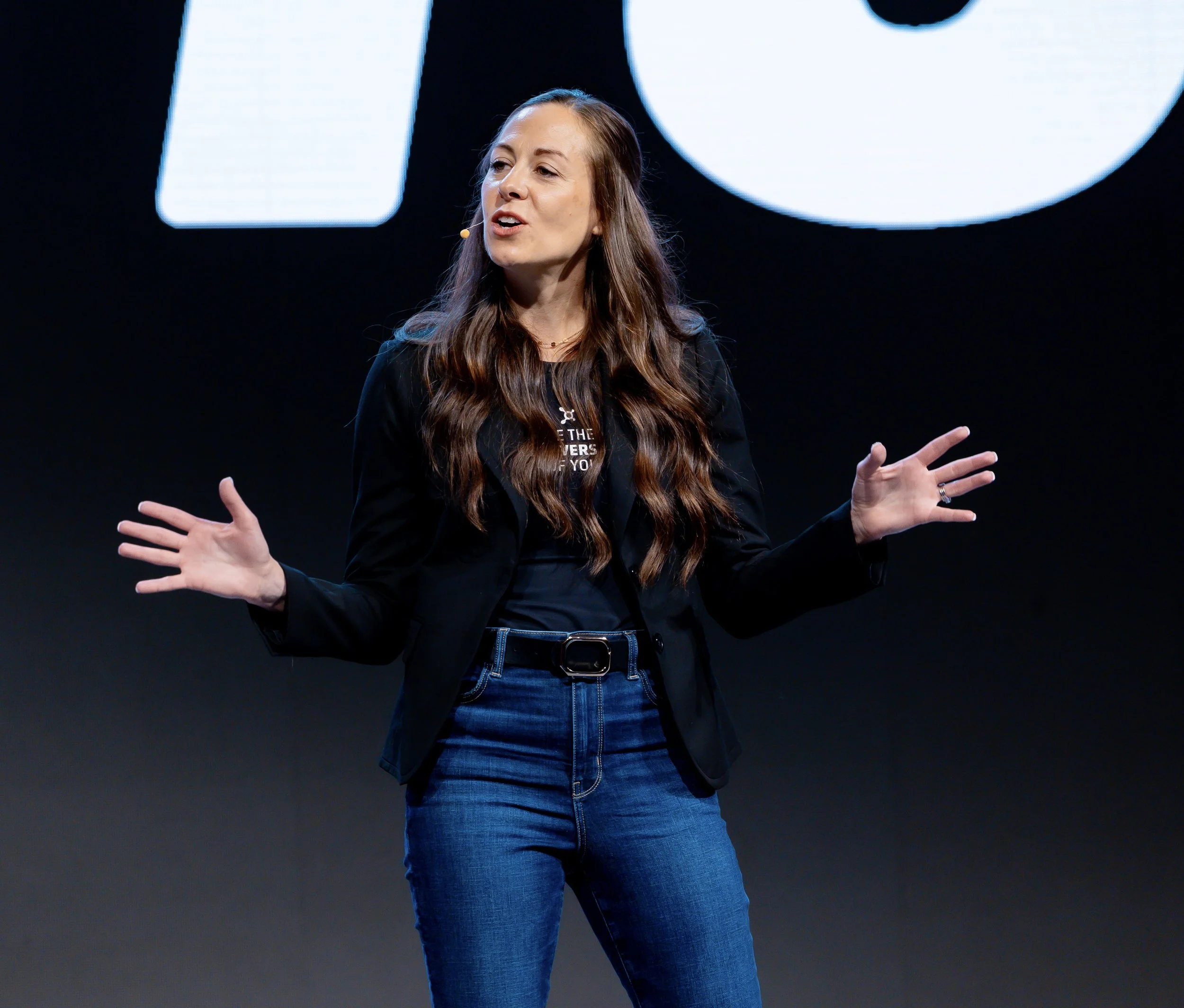 A woman with long brown hair wearing a black blazer and blue jeans speaking on stage, gesturing with her hands, standing in front of a dark background with large white letters.