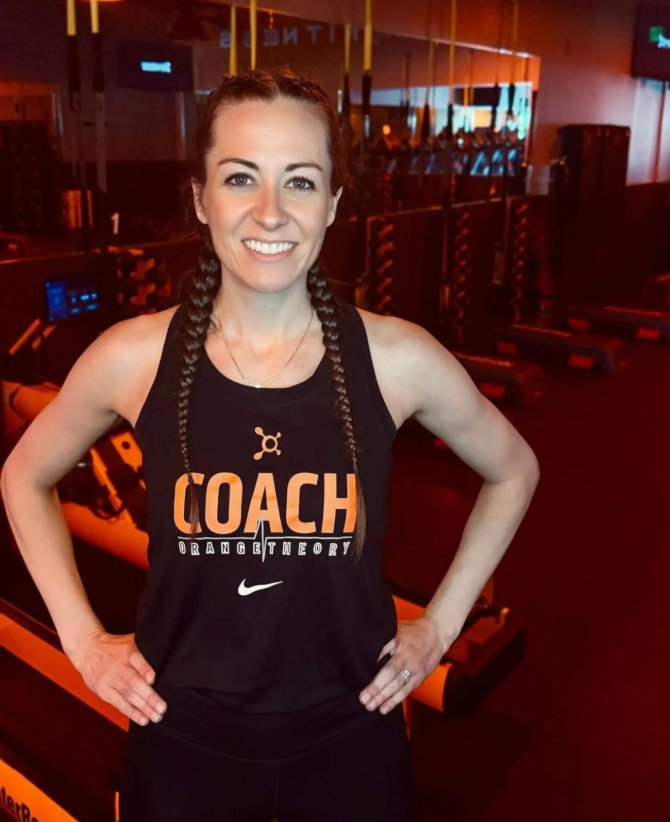A woman with braided hair smiling in a gym, wearing a black tank top with Orange Theory Coach logo, with workout equipment and orange lighting in the background.