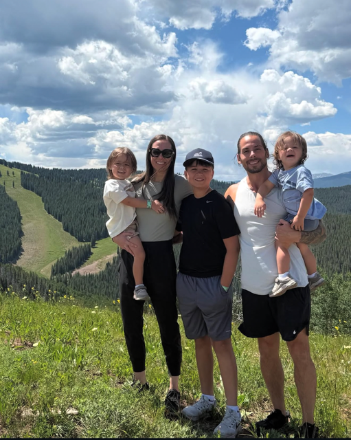 Family of five standing outdoors in a grassy field with mountains and a partly cloudy sky in the background.