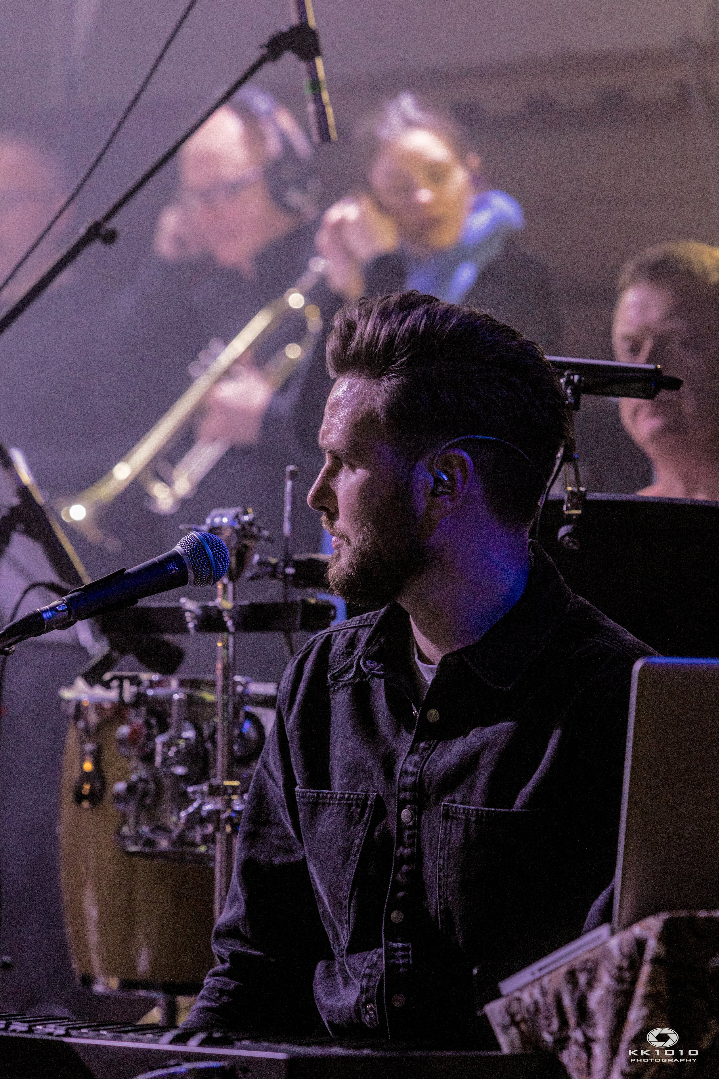 A man with dark hair and beard in profile, sitting at a keyboard with a microphone, with a group of musicians behind him, including a woman holding a guitar and a woman in the background with headphones.