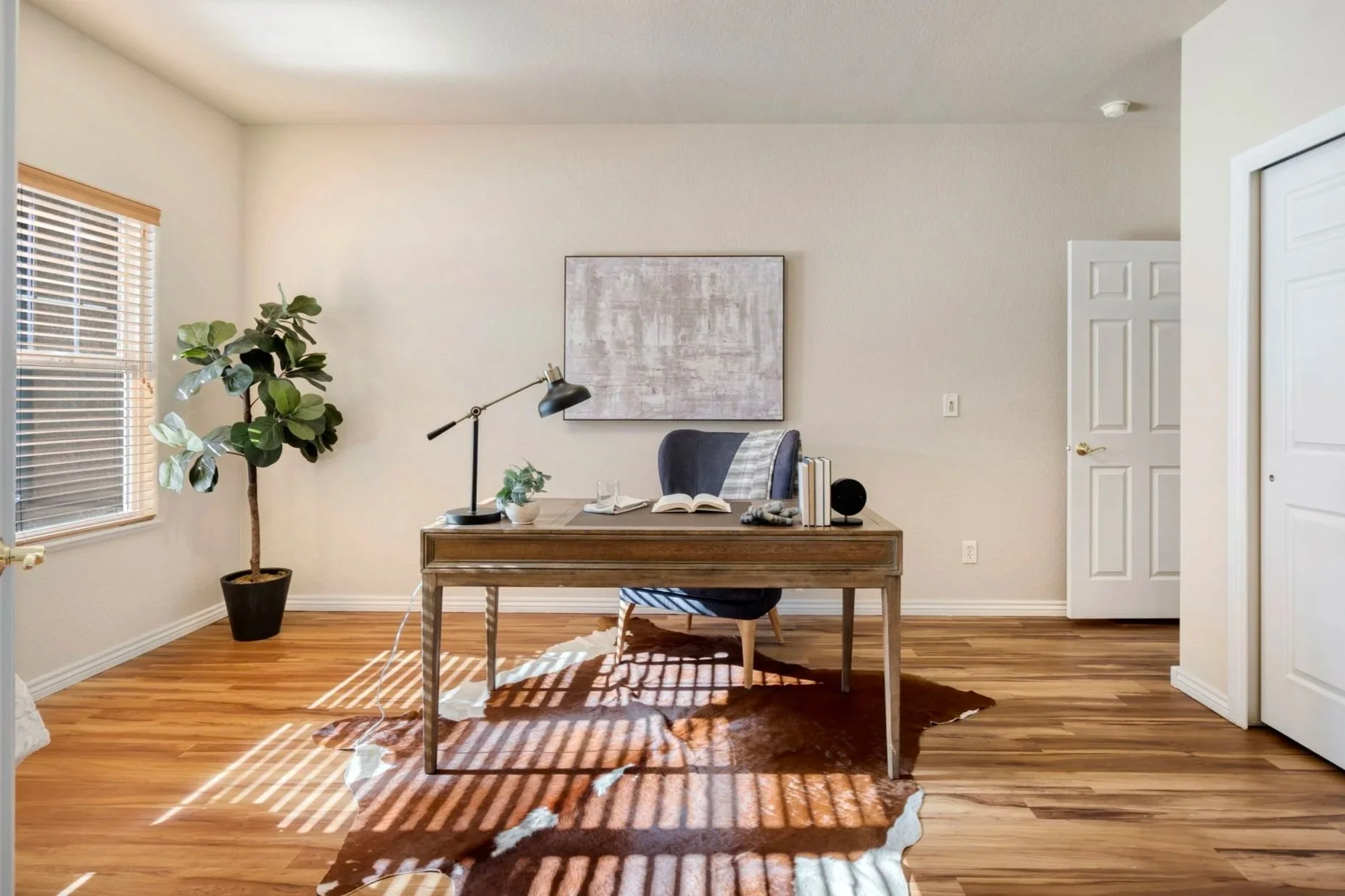 Home office staged with wooden desk, black desk lamp, open book, a blue chair, potted plant, large window with blinds, framed abstract art on the wall, wooden flooring with cowhide rug, closed white closet door.