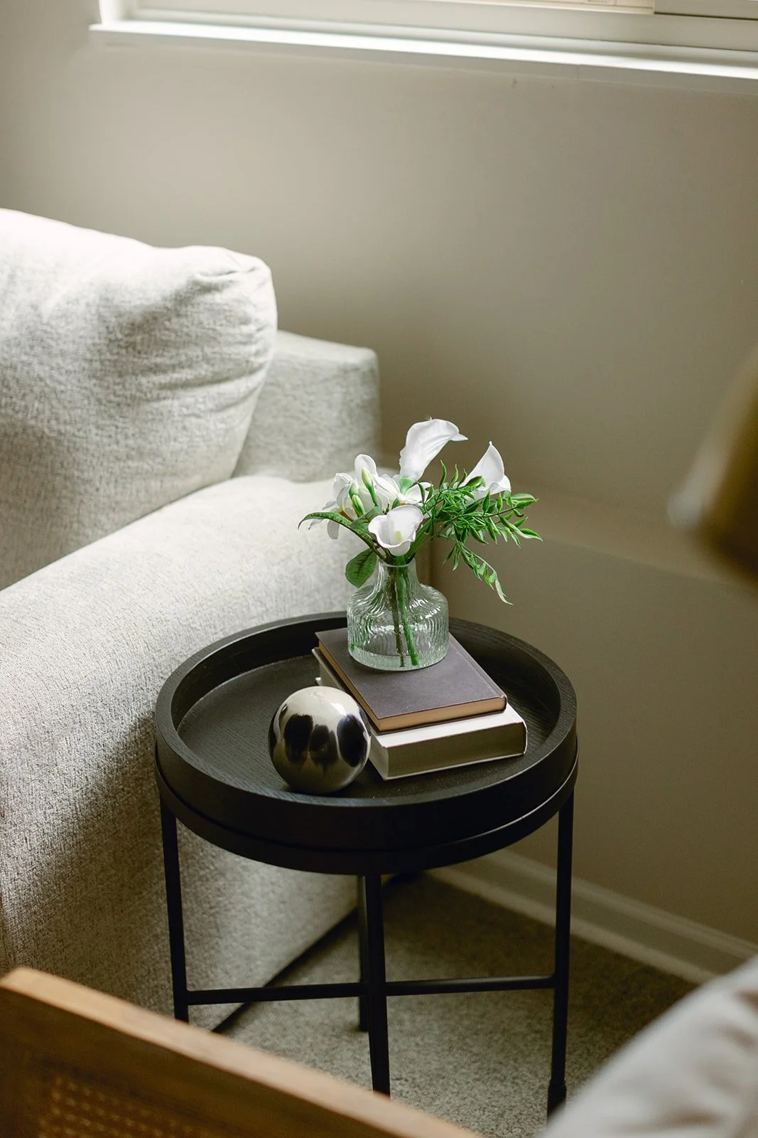A staged cozy living room corner featuring a round black side table with a glass vase of white flowers, two stacked books, and a metallic decorative ball, next to a beige couch and a window.
