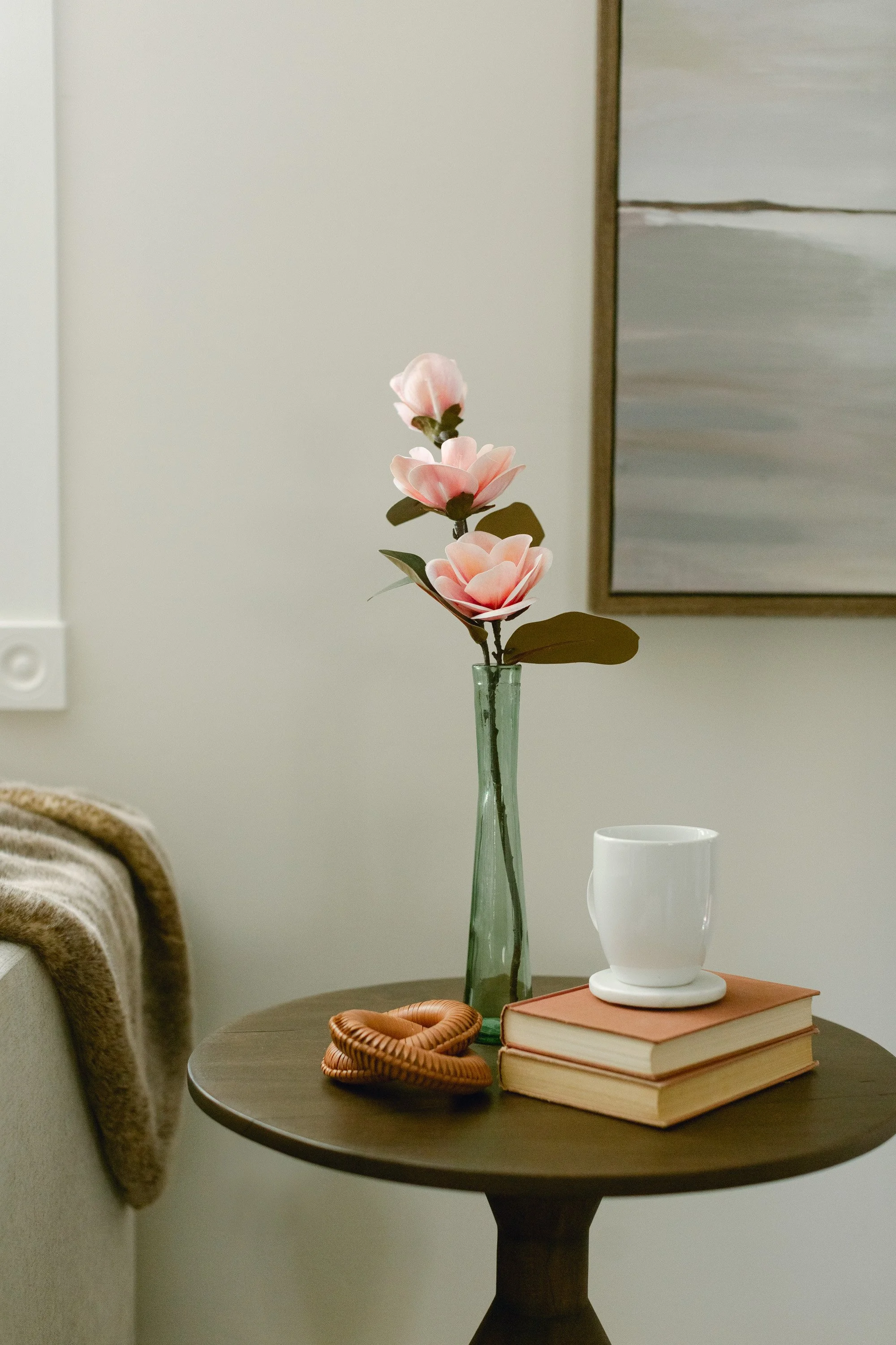 A small round wooden table staged with a vase of pink flowers, a white coffee mug, two stacked books, and a woven bracelet, next to a sofa with a blanket, and a wall with abstract art.
