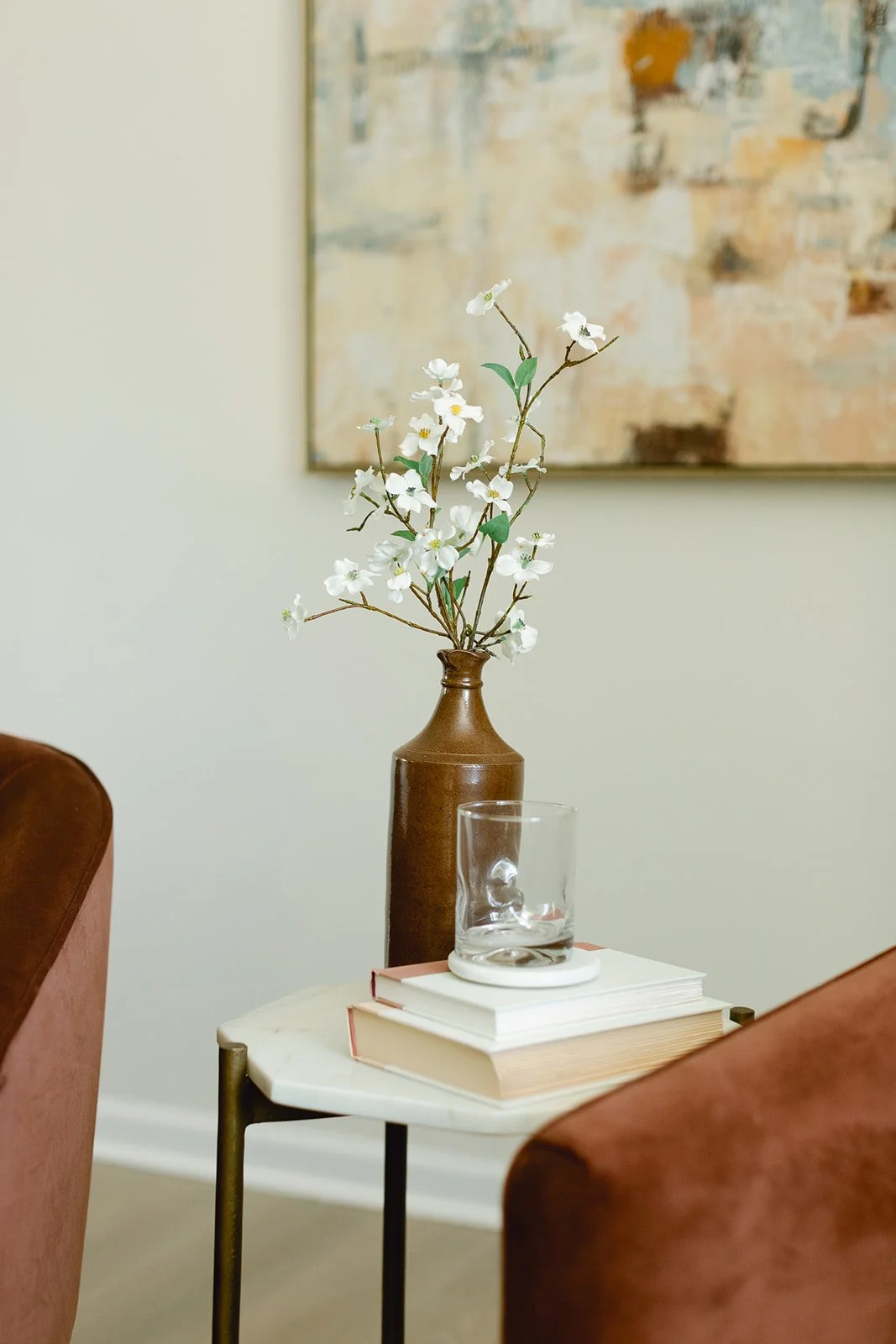 A elegantly staged small round side table with a botanical print on the wall behind it. On the table, there is a brown ceramic vase with white flowers, a stack of three books, and a clear glass placed on a white coaster.
