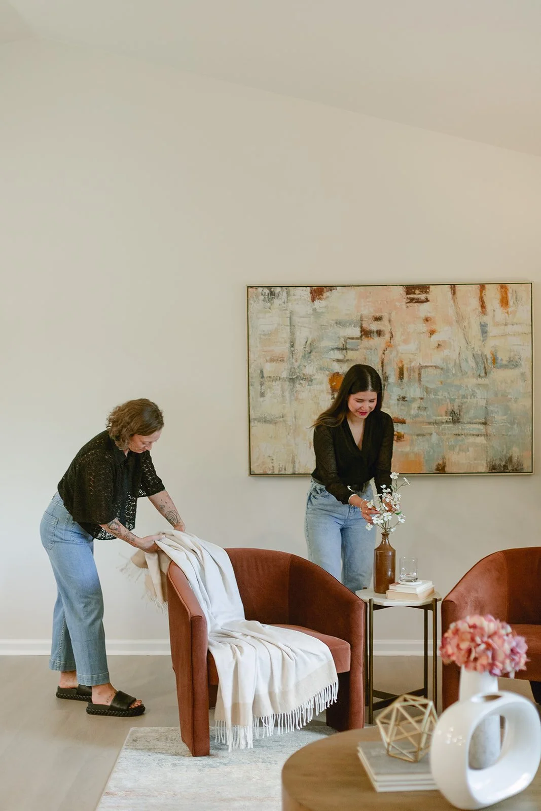 Two Modern Interior Staging Co. stagers are preparing a cozy living room, one folding a blanket on a rusty pink armchair and the other arranging flowers in a brown vase on a small side table, with abstract artwork on the wall behind them.