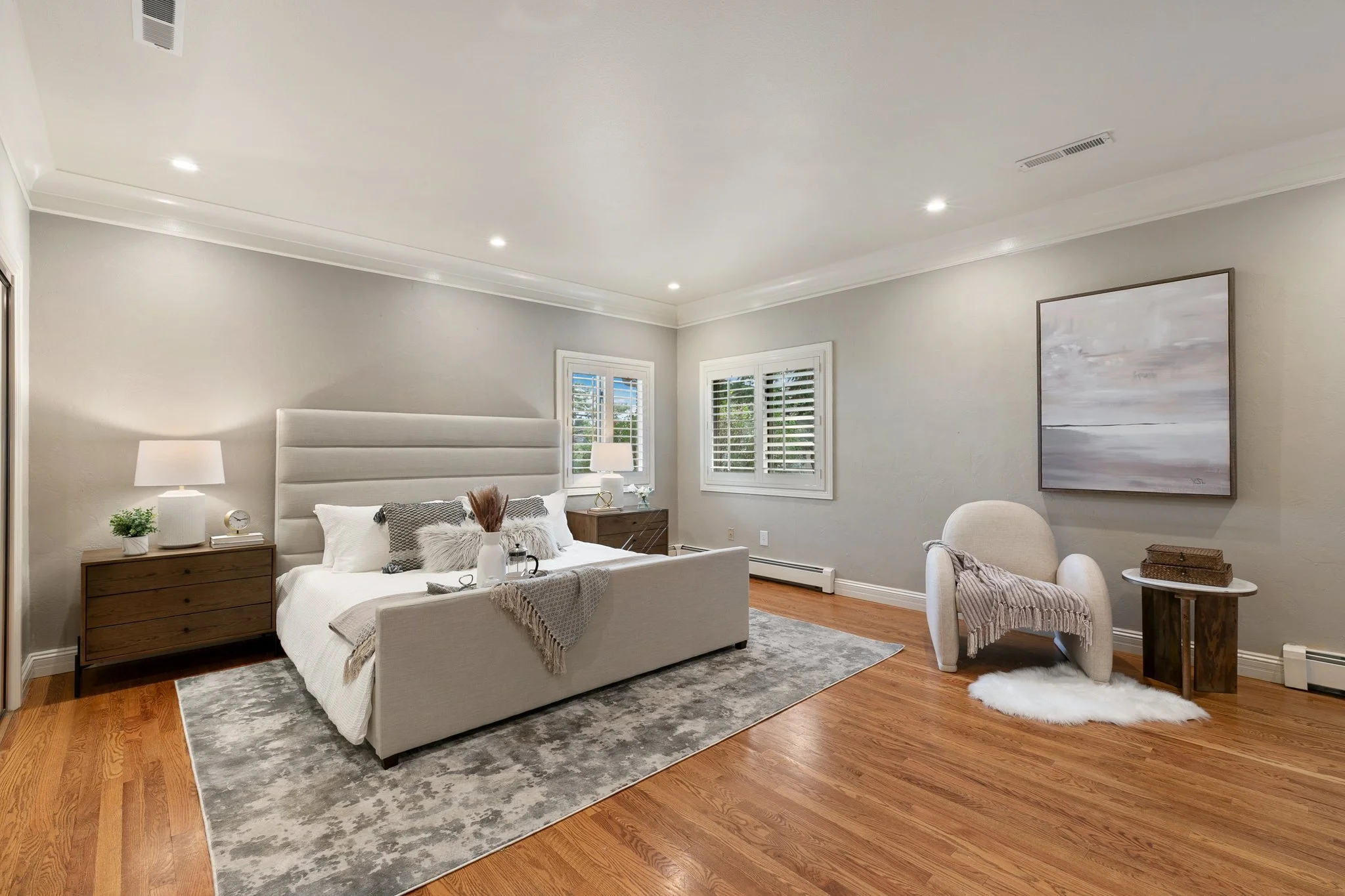 Modern bedroom staged with a large bed, two nightstands with lamps, a textured rug, a cozy armchair with a throw blanket, hardwood floors, and minimalist decor.