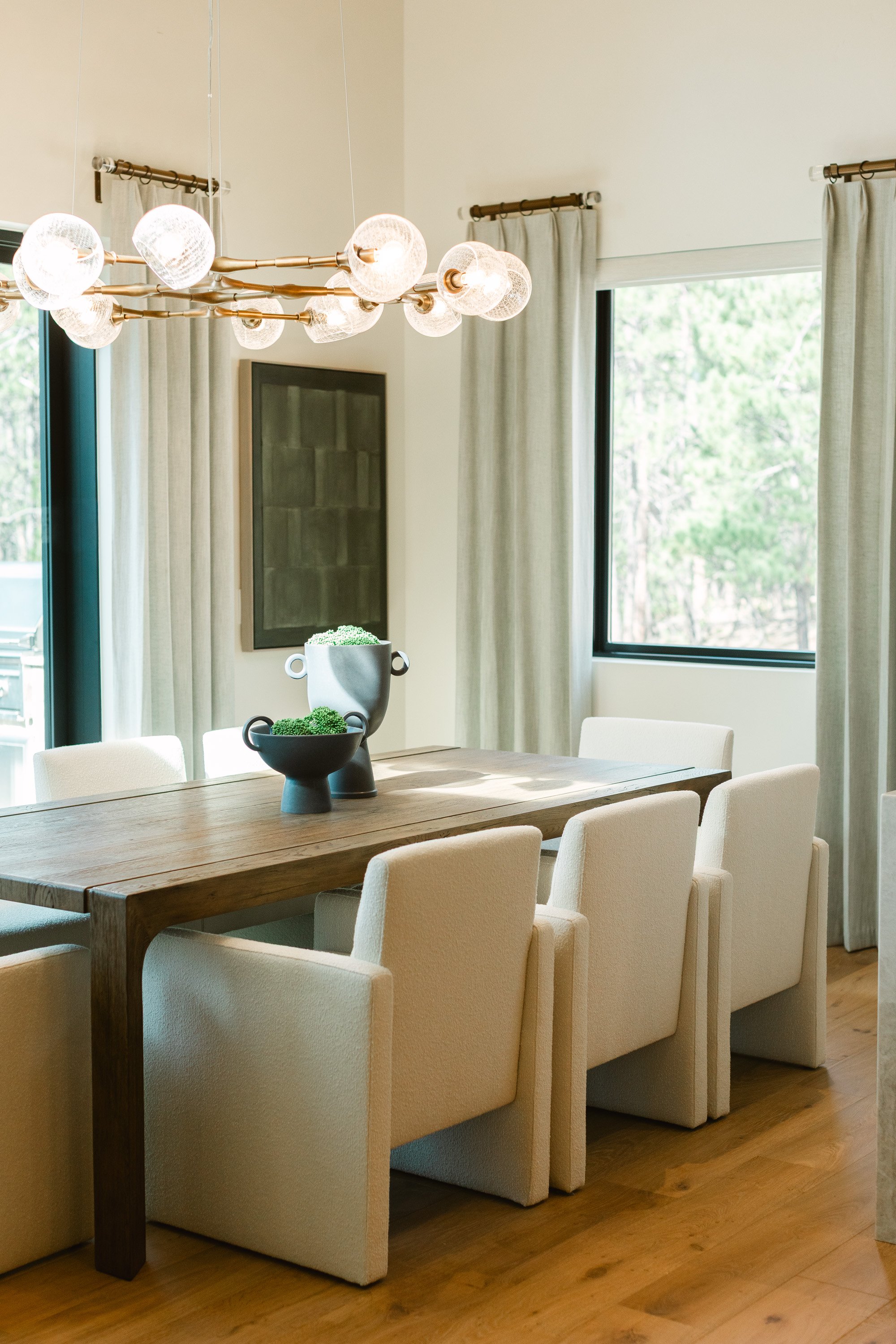 A dining room staged with a wooden table, six beige upholstered chairs, a modern chandelier with multiple glass bulbs, large windows with beige curtains, a wall art piece, and two decorative vases with green plants on the table.