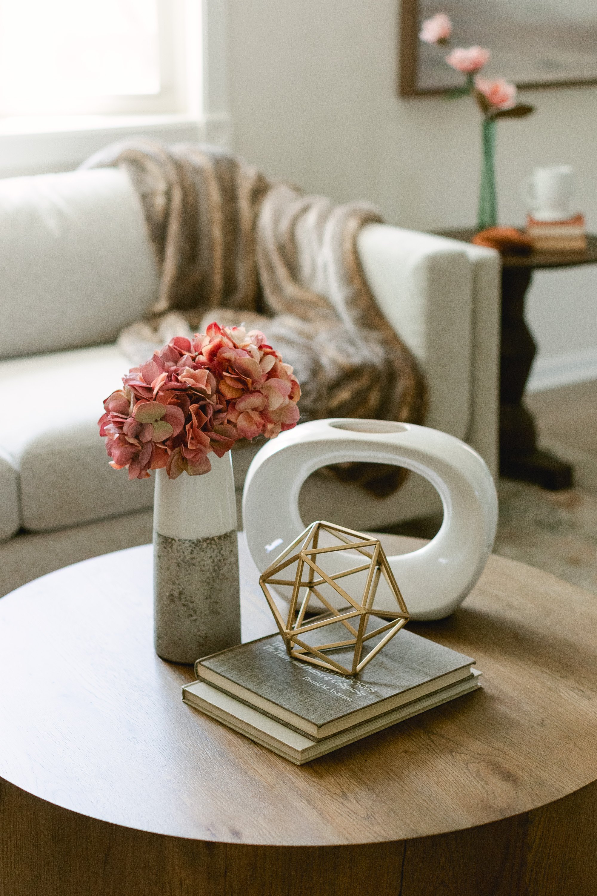 A living room staged with a wooden coffee table, a floral arrangement, geometric sculpture, and books, with a sofa, blanket, side table with a vase, and a window in the background.