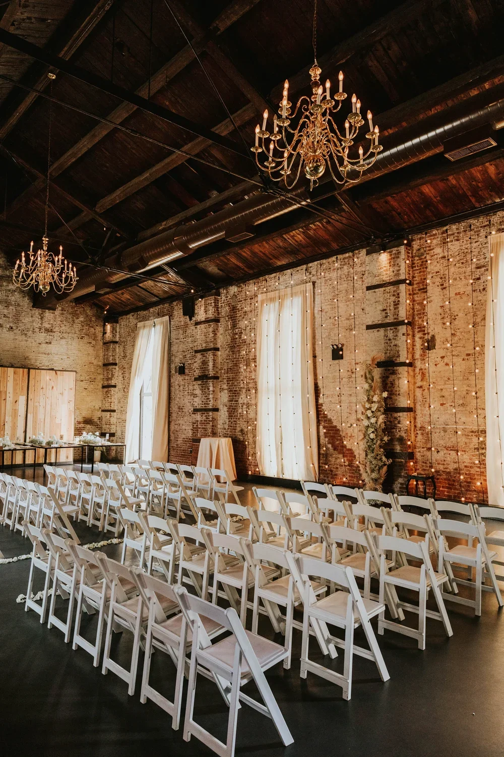 Elegant wedding or event venue with rows of white chairs, rustic brick walls, large windows with white curtains, chandeliers, and decorative hanging lights.