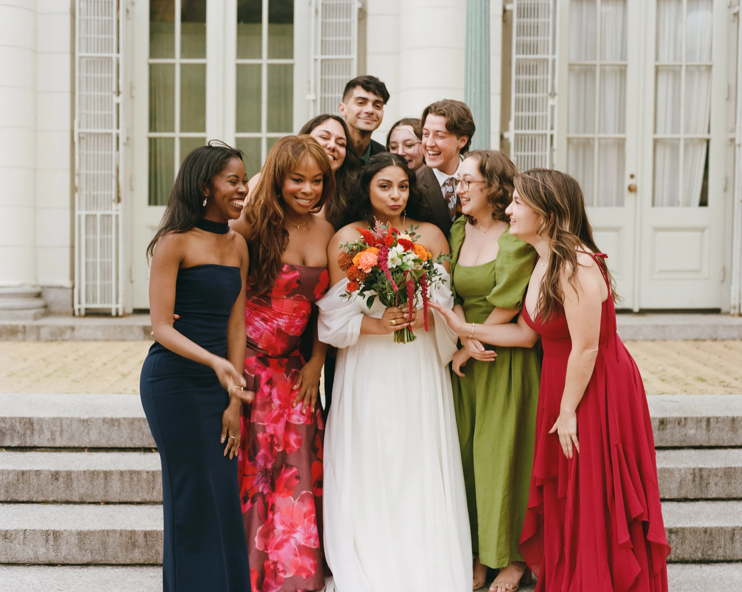 A group of people gathered on steps in front of a building, celebrating with a woman holding a colorful bouquet of flowers, dressed in a white wedding gown. The group appears joyful and happy.