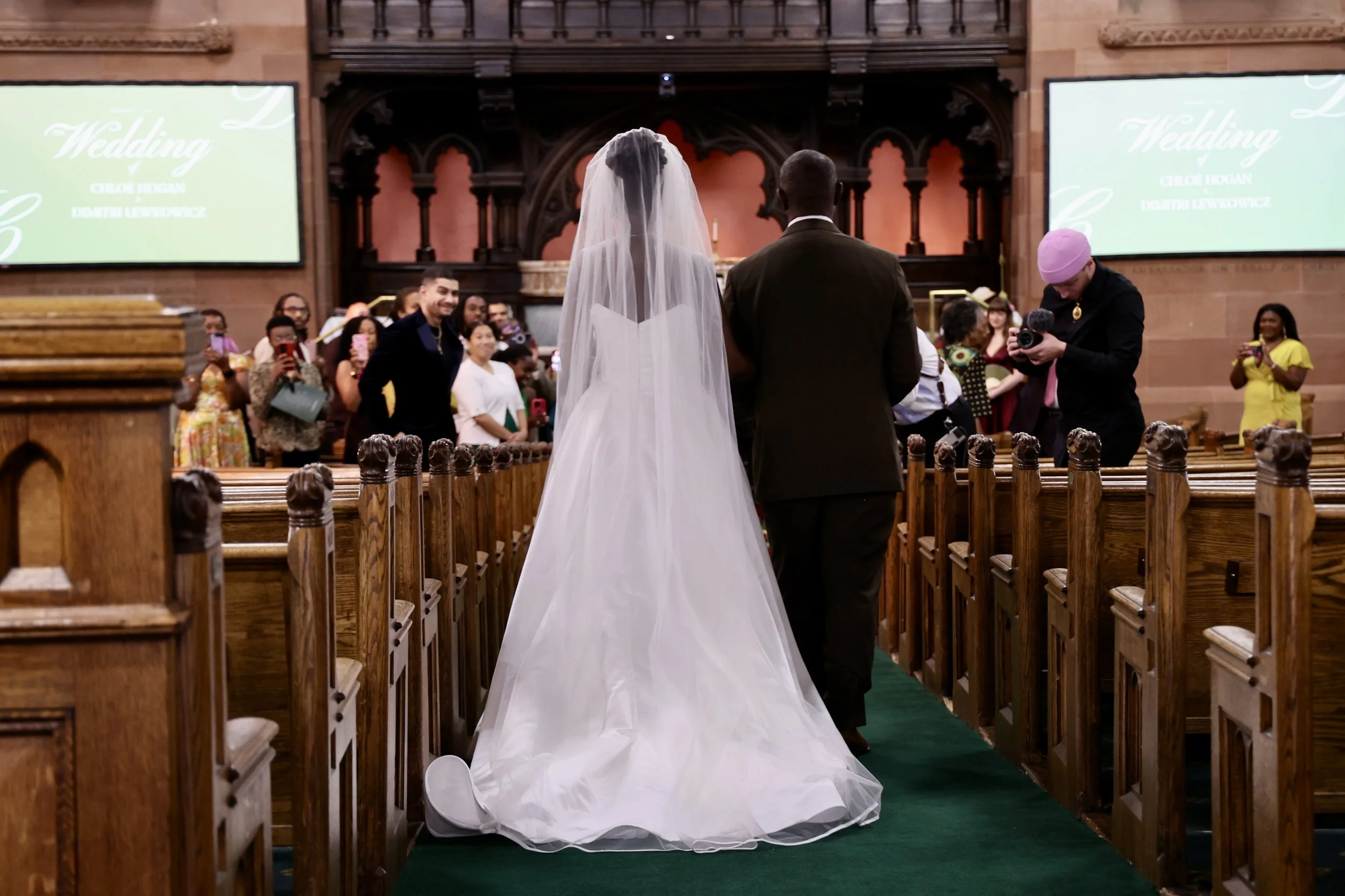 Bride and groom standing at the altar during a wedding ceremony inside a church, with guests seated and taking photos, and two large screens displaying wedding information.