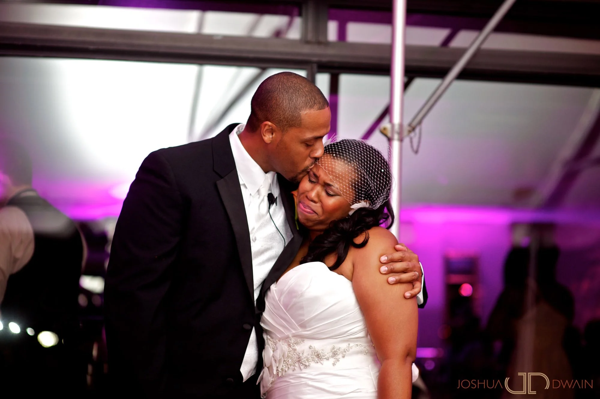 A man in a tuxedo kisses a woman in a wedding dress on the forehead, hugging her, during a wedding celebration.