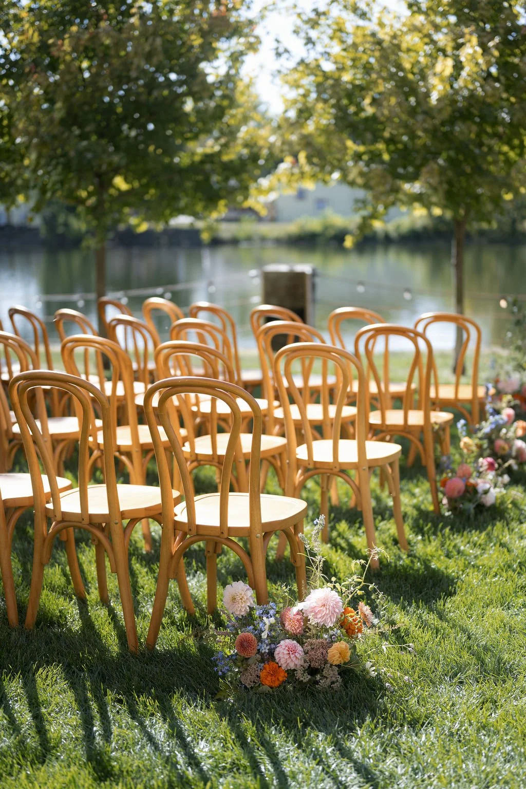 Outdoor wedding ceremony setup with wooden chairs arranged in front of a lake, decorated with colorful flowers and surrounded by trees.