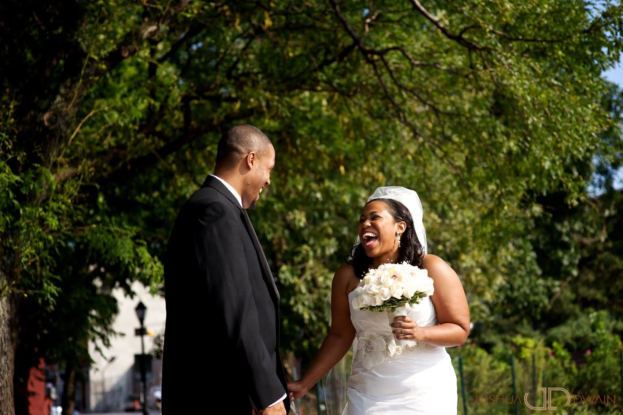 A bride and groom standing outdoors under a large green leafy tree, smiling and holding hands during their wedding ceremony.