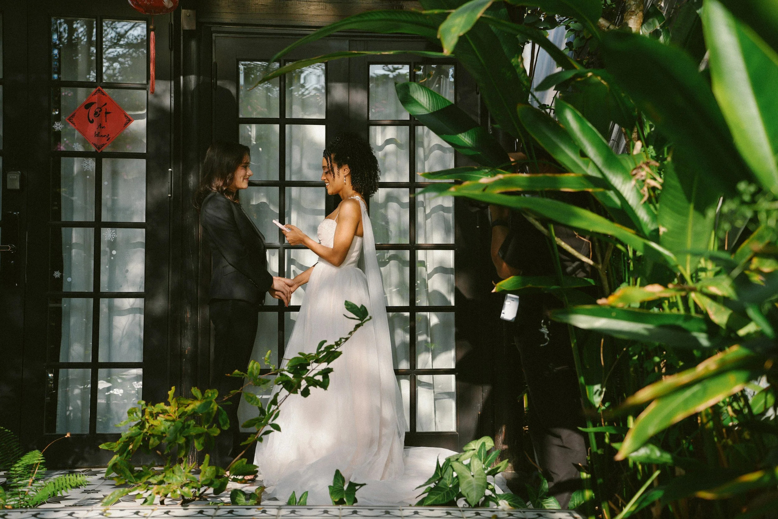 Two women, one in a black suit and the other in a white dress, hold hands and smile at each other in front of black windowed doors with a red Chinese New Year decoration and hanging snowflake stickers, surrounded by lush green plants.