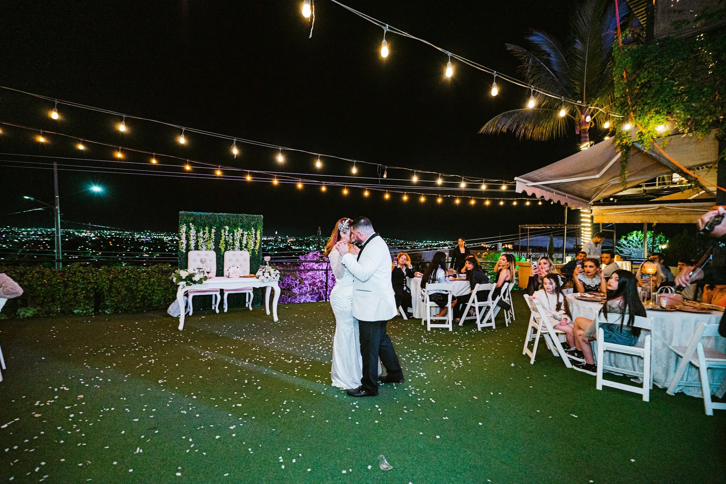 Nighttime outdoor wedding celebration with couples dancing, guest tables, string lights, and city skyline in the background.