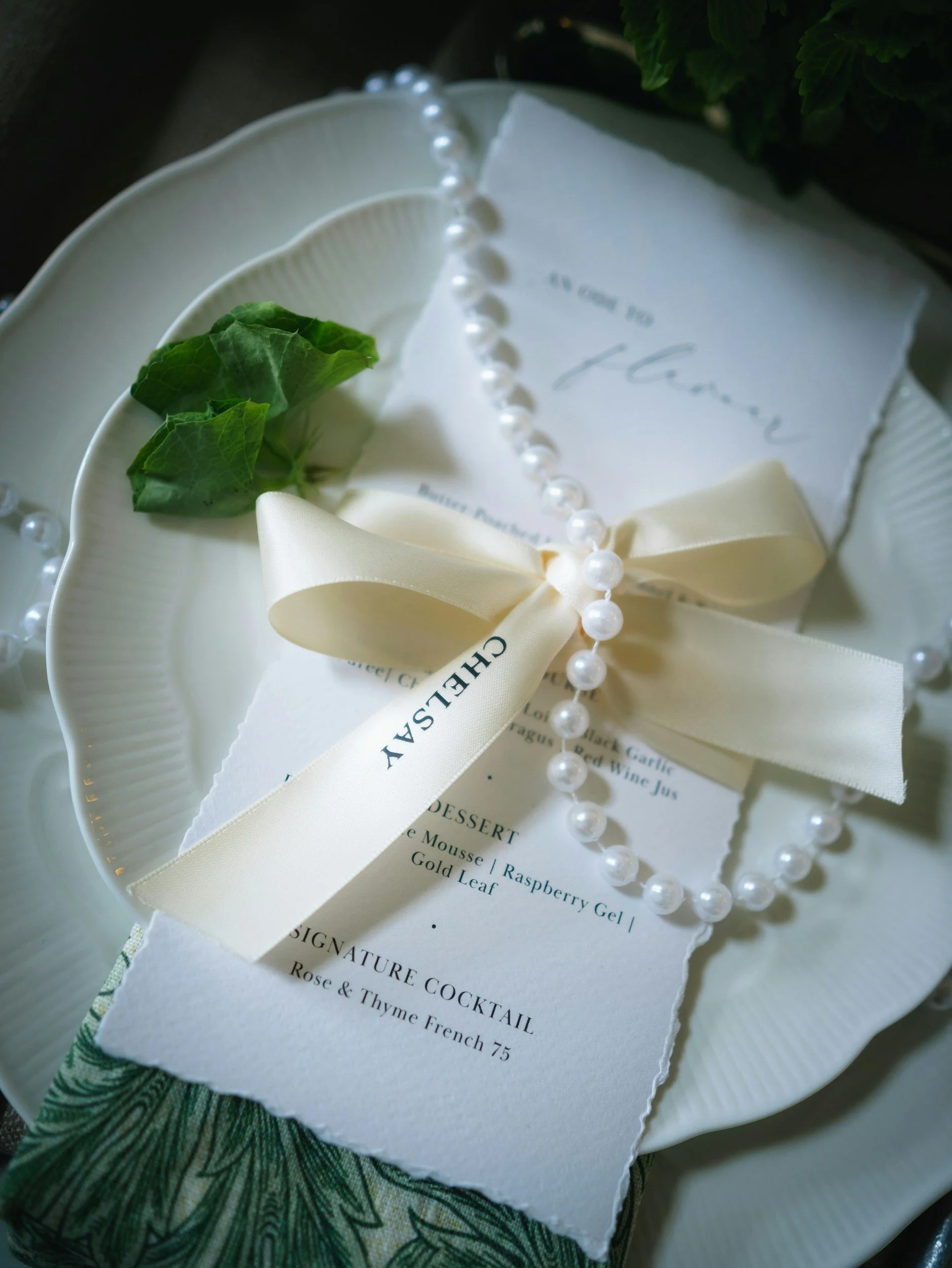 A close-up of a white plate with a menu, a string of pearl beads, a cream-colored ribbon, and a small sprig of green leaves placed on top.
