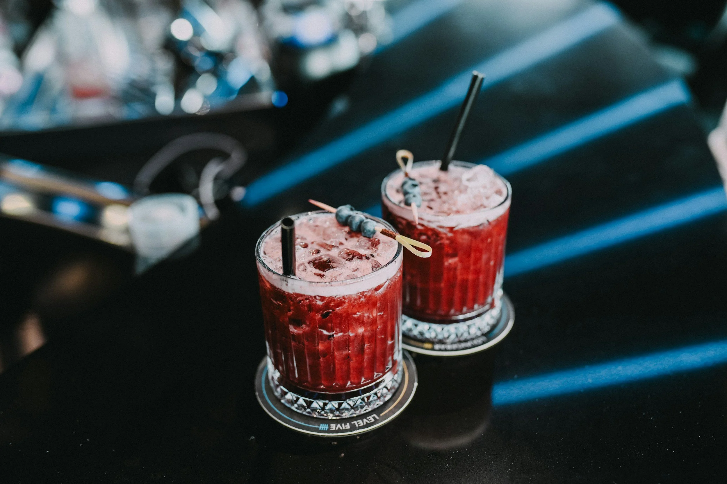Two red beverages with ice and blueberries in tall glasses on a black surface.
