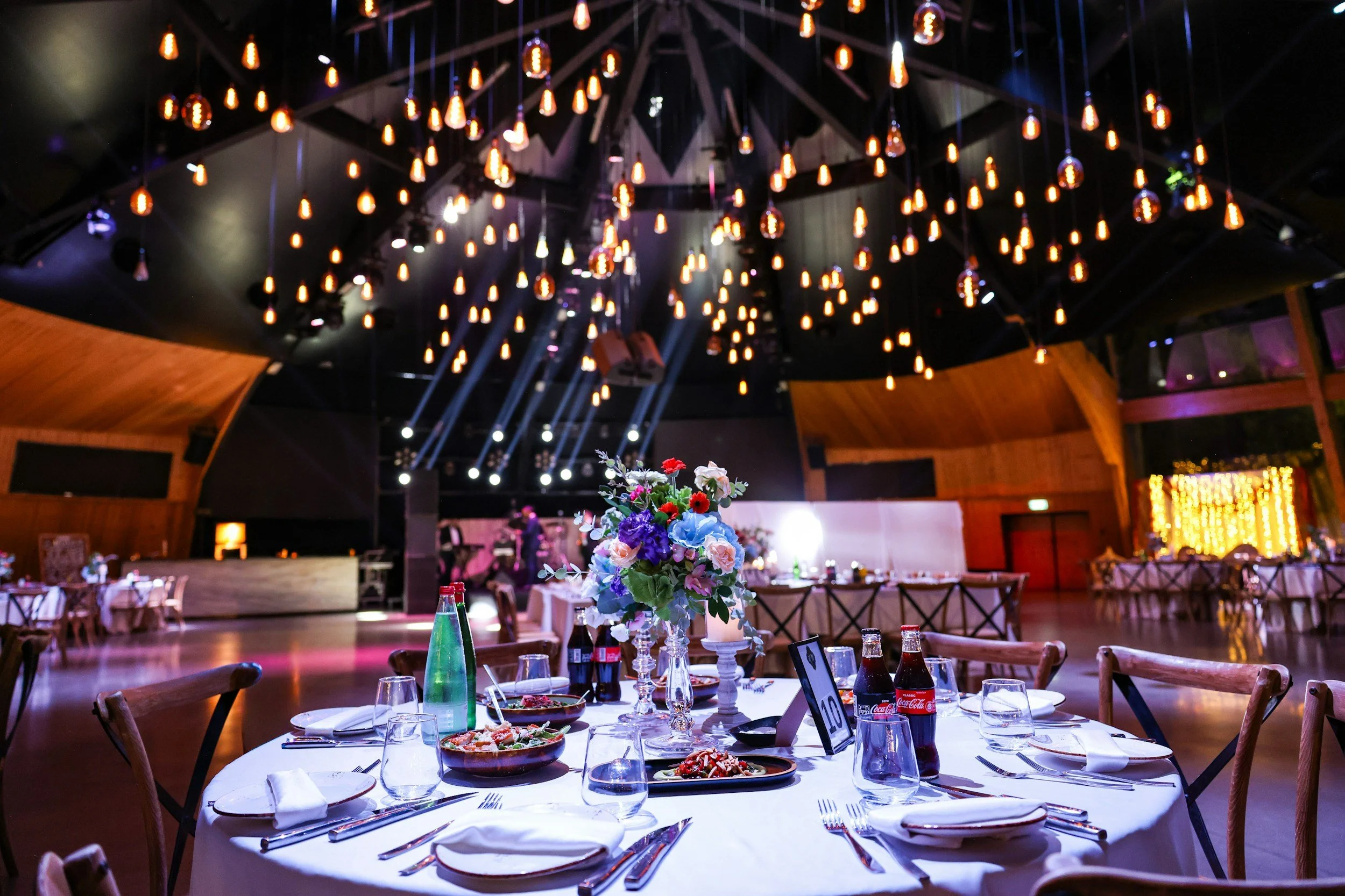 A banquet hall with round tables set for a meal, decorated with white tablecloths, flowers, and drinks, and illuminated by an elaborate chandelier with hanging lights.