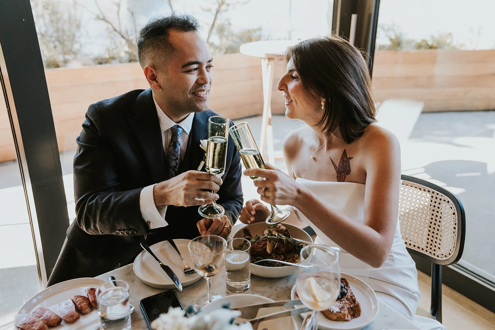 A man in a suit and a woman in a white dress toast with champagne glasses at a dinner table, smiling at each other, with food on the table and a sunlit window in the background.