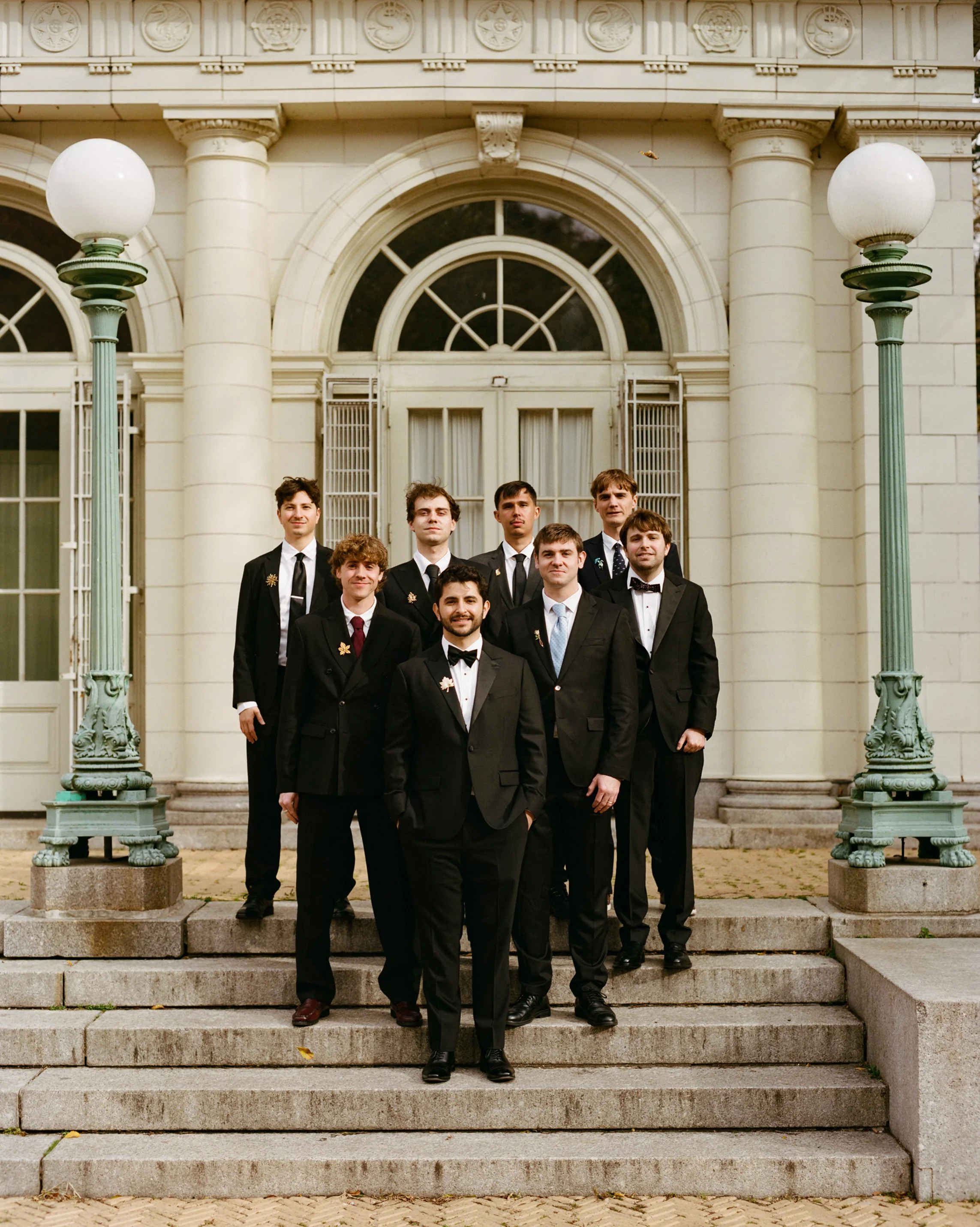 A group of young men dressed in black tuxedos with bow ties, standing on steps outside a historic white building with large windows and columns, posing for a formal photo.