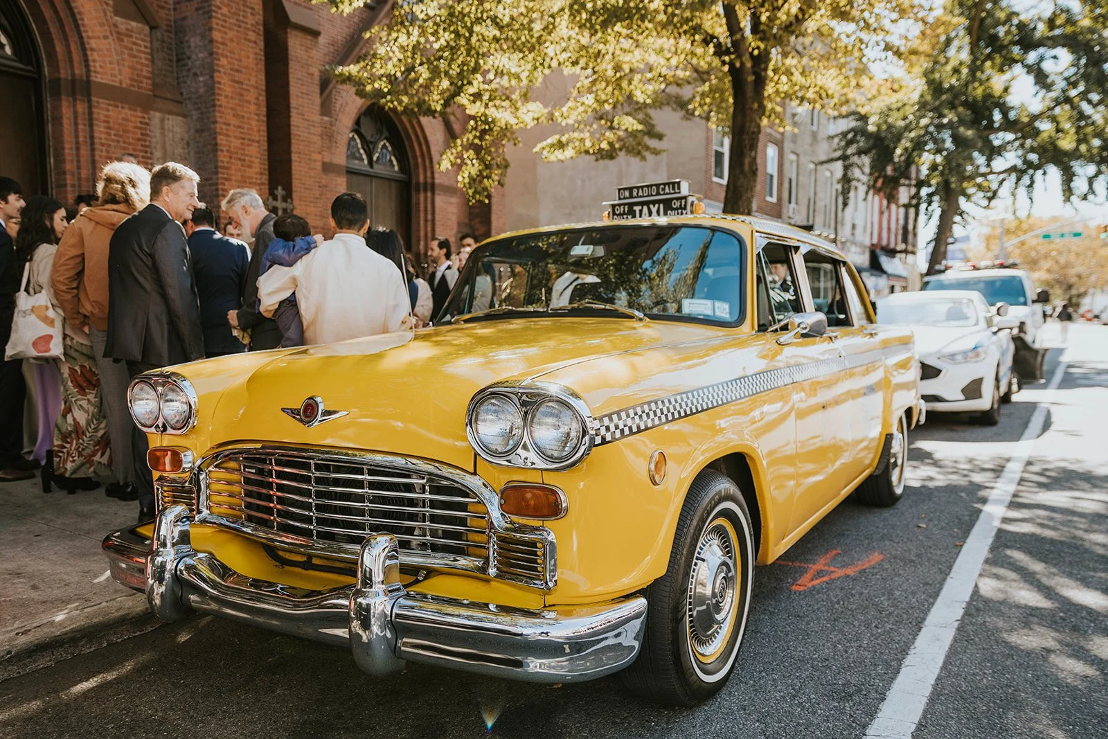 A yellow vintage taxi cab parked on a city street with a crowded group of people gathered nearby on the sidewalk, trees with green leaves overhead, and other cars in the background.