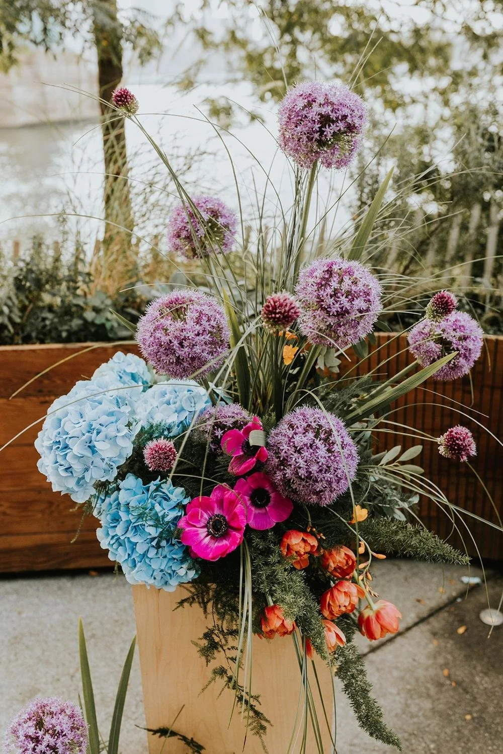 Colorful flower arrangement with purple, blue, pink, and orange flowers in a wooden vase outdoors.