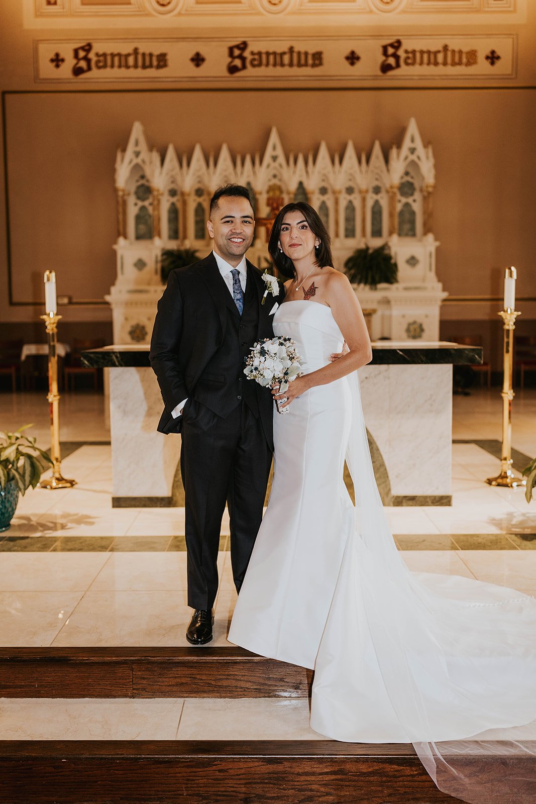 A bride and groom standing together inside a church. The bride is wearing a white wedding gown and holding a bouquet of white flowers, while the groom is dressed in a black suit. The background features an altar with a Gothic-style backdrop and two t