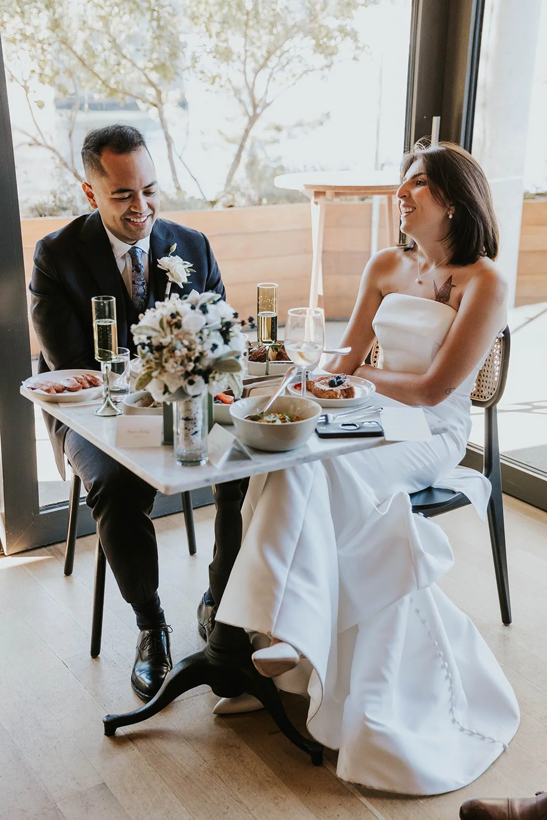 A bride and groom sitting at a table during their wedding celebration, smiling and enjoying a meal together.