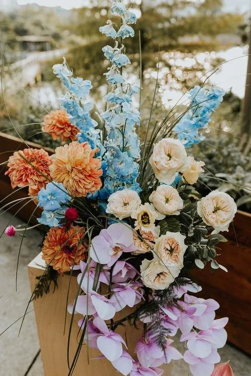 A colorful bouquet of flowers including blue delphiniums, peach dahlias, white roses, and pink orchids arranged in a wooden vase outdoors.