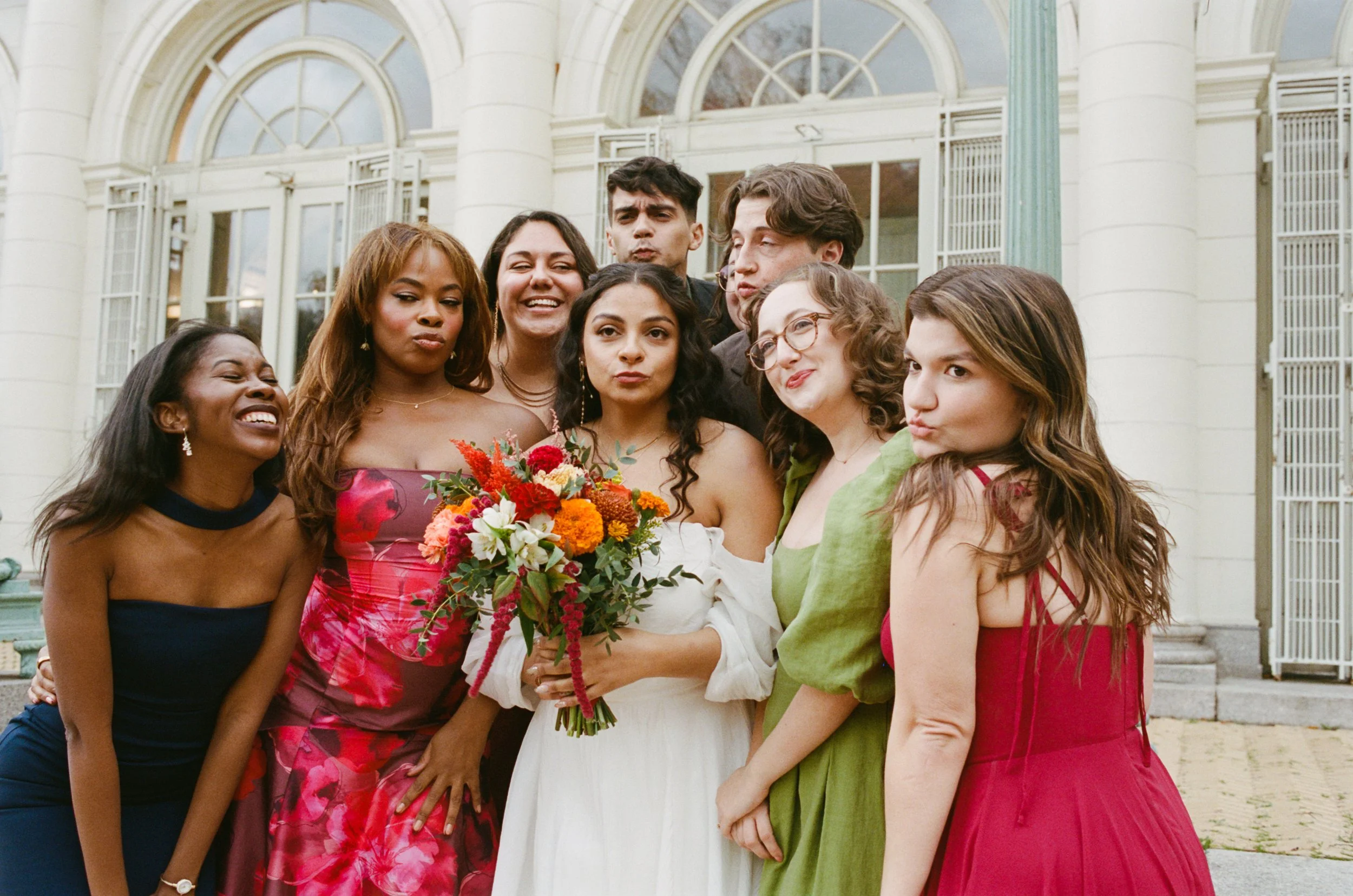 A group of diverse friends gathered outside a building, celebrating a wedding, with a woman holding a bouquet of colorful flowers.