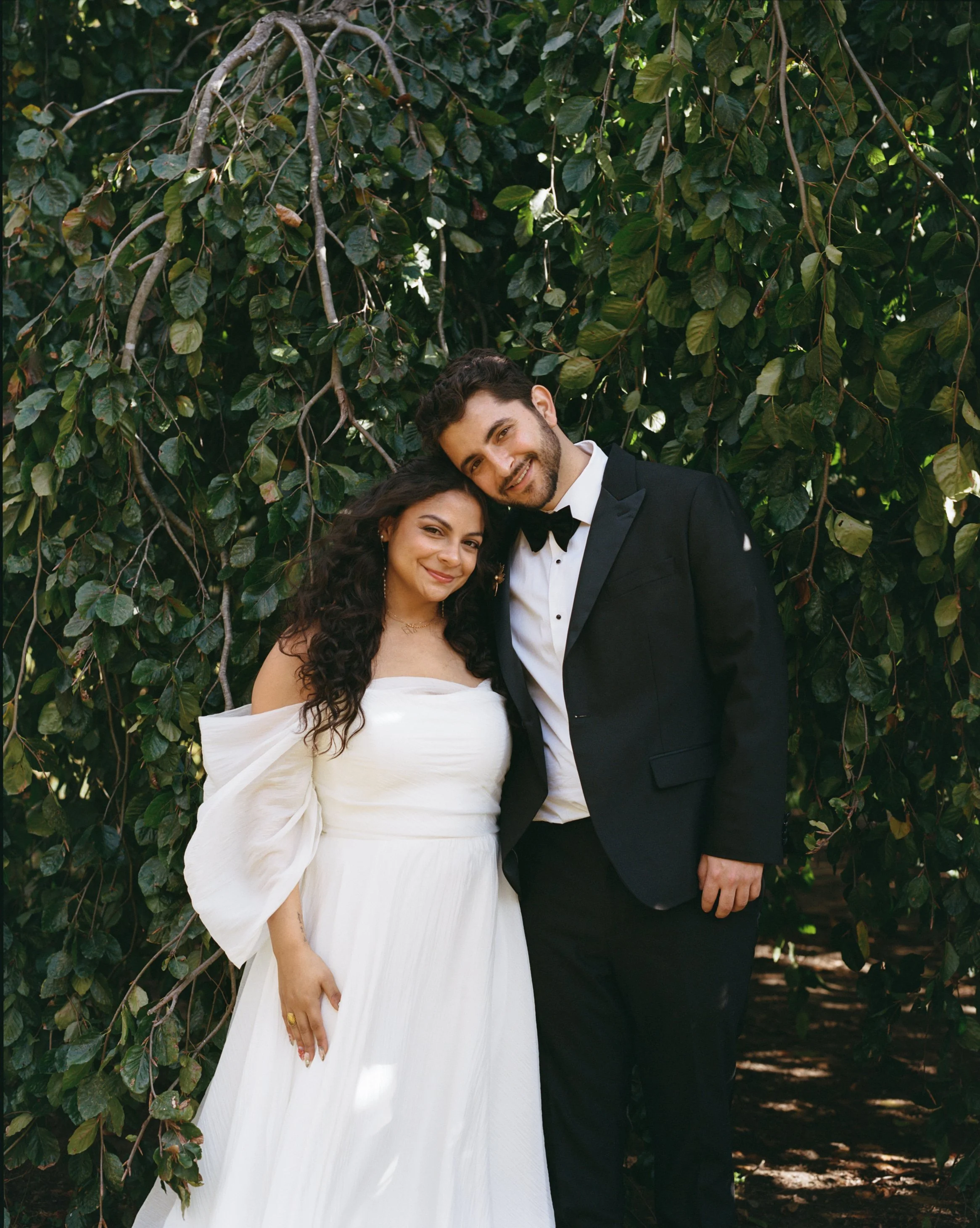 A smiling couple dressed in wedding attire standing in front of a lush, green leafy bush. The woman is wearing an off-the-shoulder white wedding gown, and the man is wearing a black tuxedo with a bow tie.